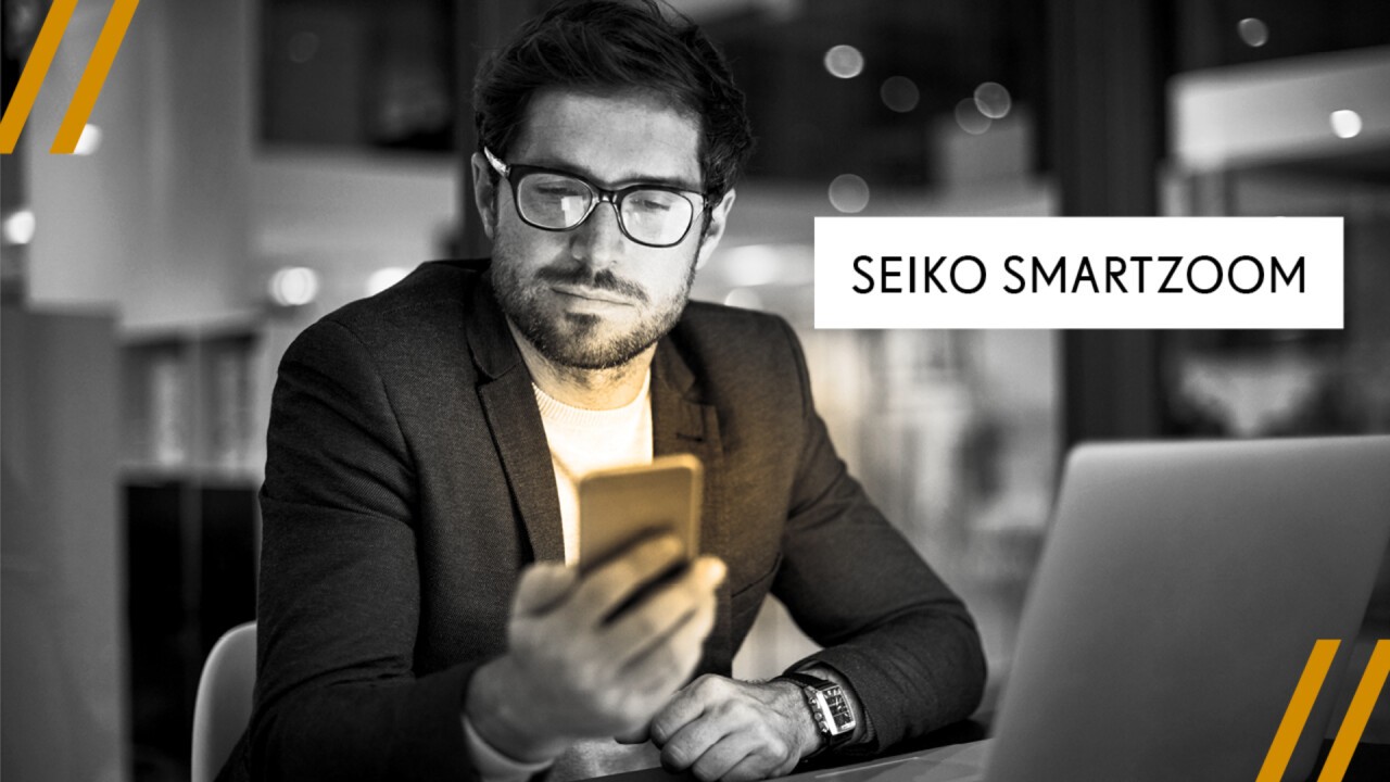Das Bild zeigt einen jüngeren männlichen Brillenträger bei der Nutzung digitaler Geräte. Es beinhaltet den Schriftzug Seiko SmartZoom.