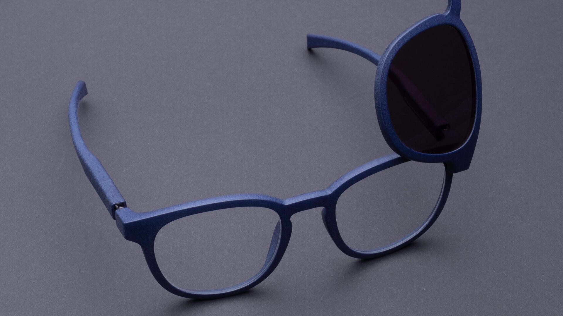 Eine blaue Brille mit einem dunklen Sonnenbrillenglas, das über das rechte Auge geklappt ist, vor einem schlichten grauen Hintergrund.