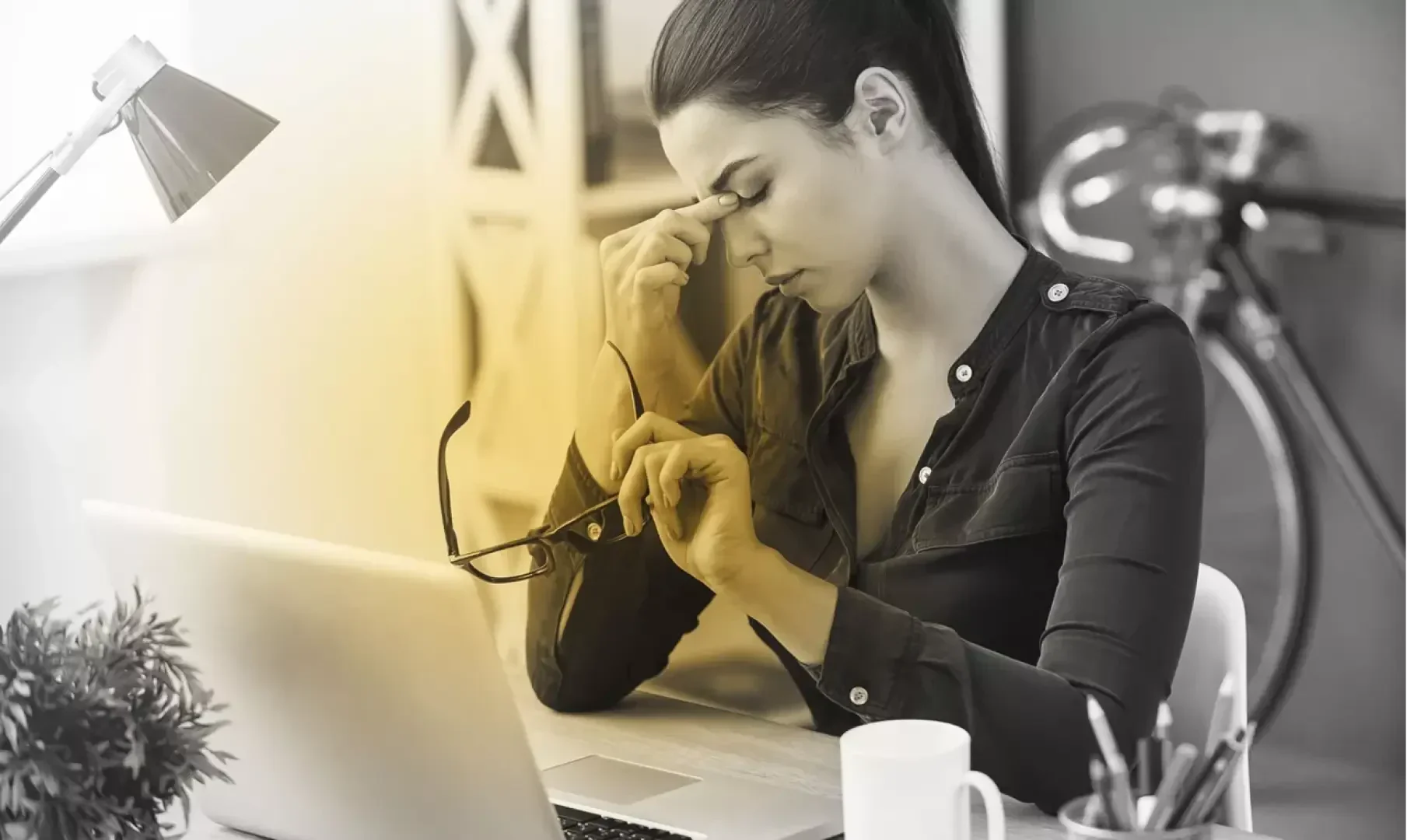 A woman sits at a desk with a laptop, holding her glasses in one hand and pinching the bridge of her nose with the other, appearing tired or stressed. A lamp, coffee mug, and plant are also on the desk.