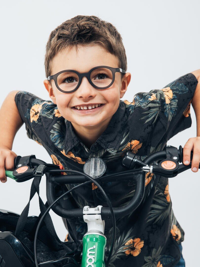 Lächelnder kleiner Junge mit kurzen braunen Haaren und Brille, der ein dunkles Hemd mit Blumenmuster trägt, den Lenker eines Fahrrads hält und vor einem schlichten hellen Hintergrund in die Kamera blickt.