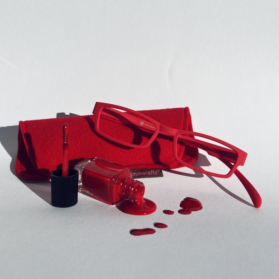 Eine Brille mit rotem Rahmen und ein dazu passendes rotes Etui liegen neben einer offenen Flasche roten Nagellacks, der auf eine weiße Oberfläche verschüttet wurde und so einen Farbtupfer erzeugt.