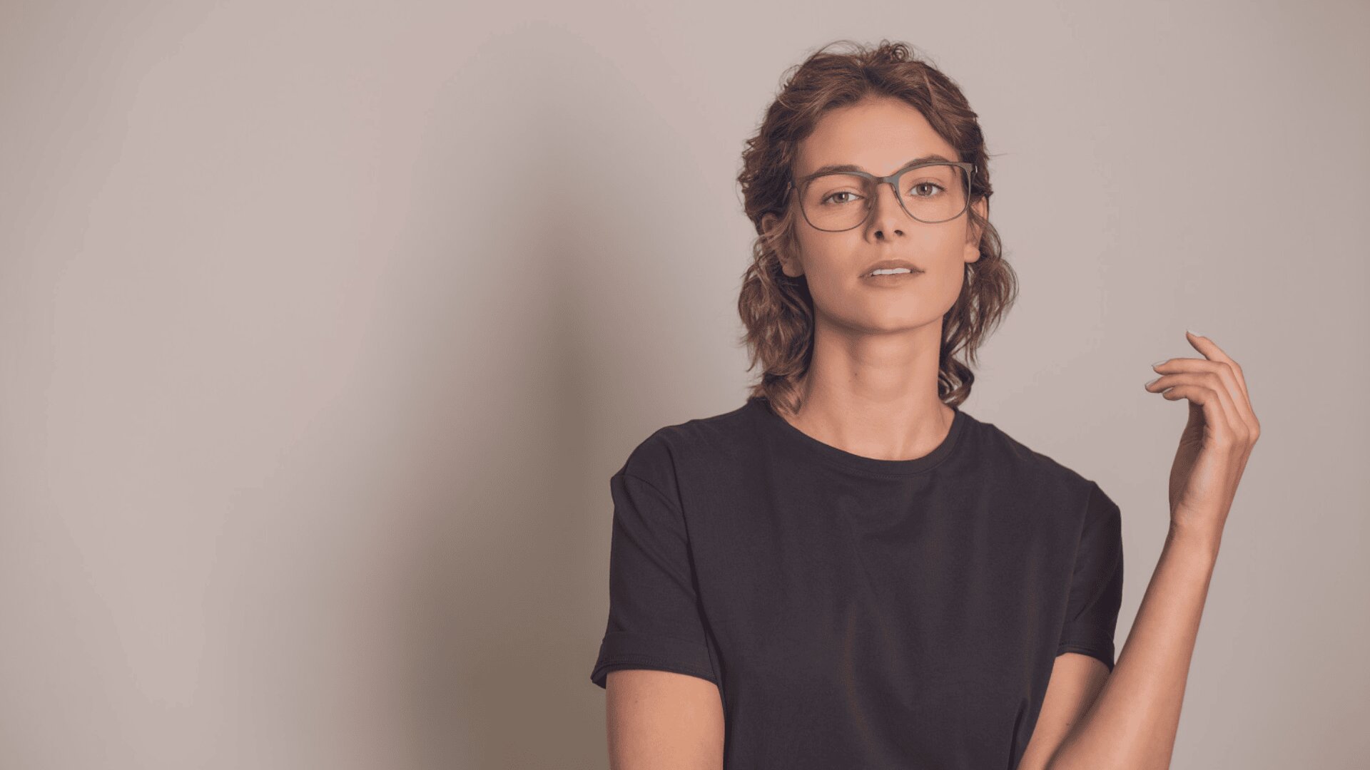 Eine Person mit kurzen, gewellten braunen Haaren, die eine Brille und ein schlichtes schwarzes T-Shirt trägt, steht vor einem hellbeigen Hintergrund und blickt mit neutraler Miene und einer erhobenen Hand in die Kamera.