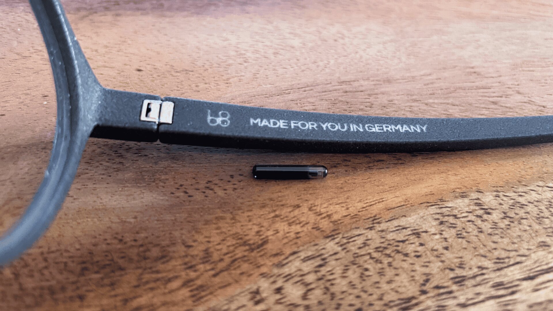 Close-up of a black eyeglass frame with text “MADE FOR YOU IN GERMANY” on the arm, next to a tiny cylindrical black microchip placed on a wooden surface.