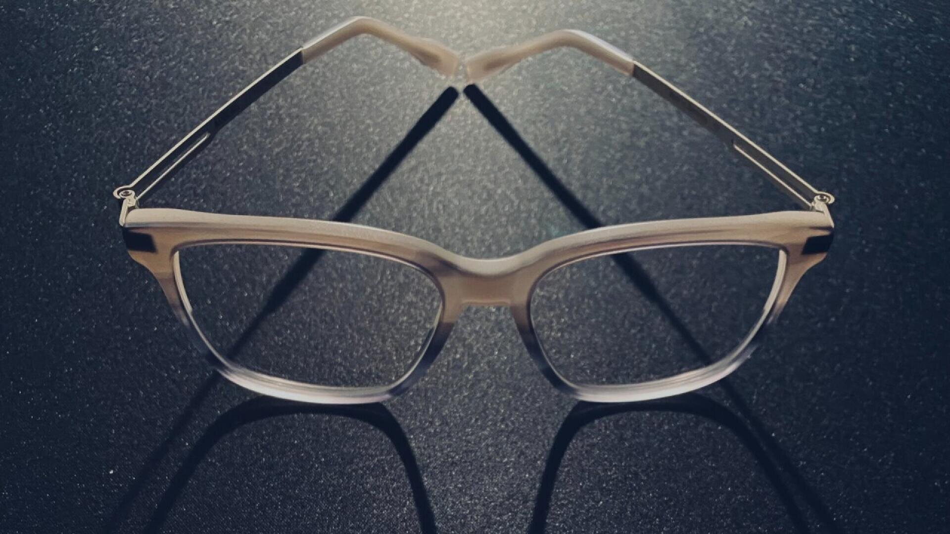 A pair of eyeglasses with clear frames rests on a dark, textured surface, casting distinct shadows that form an interesting geometric pattern.