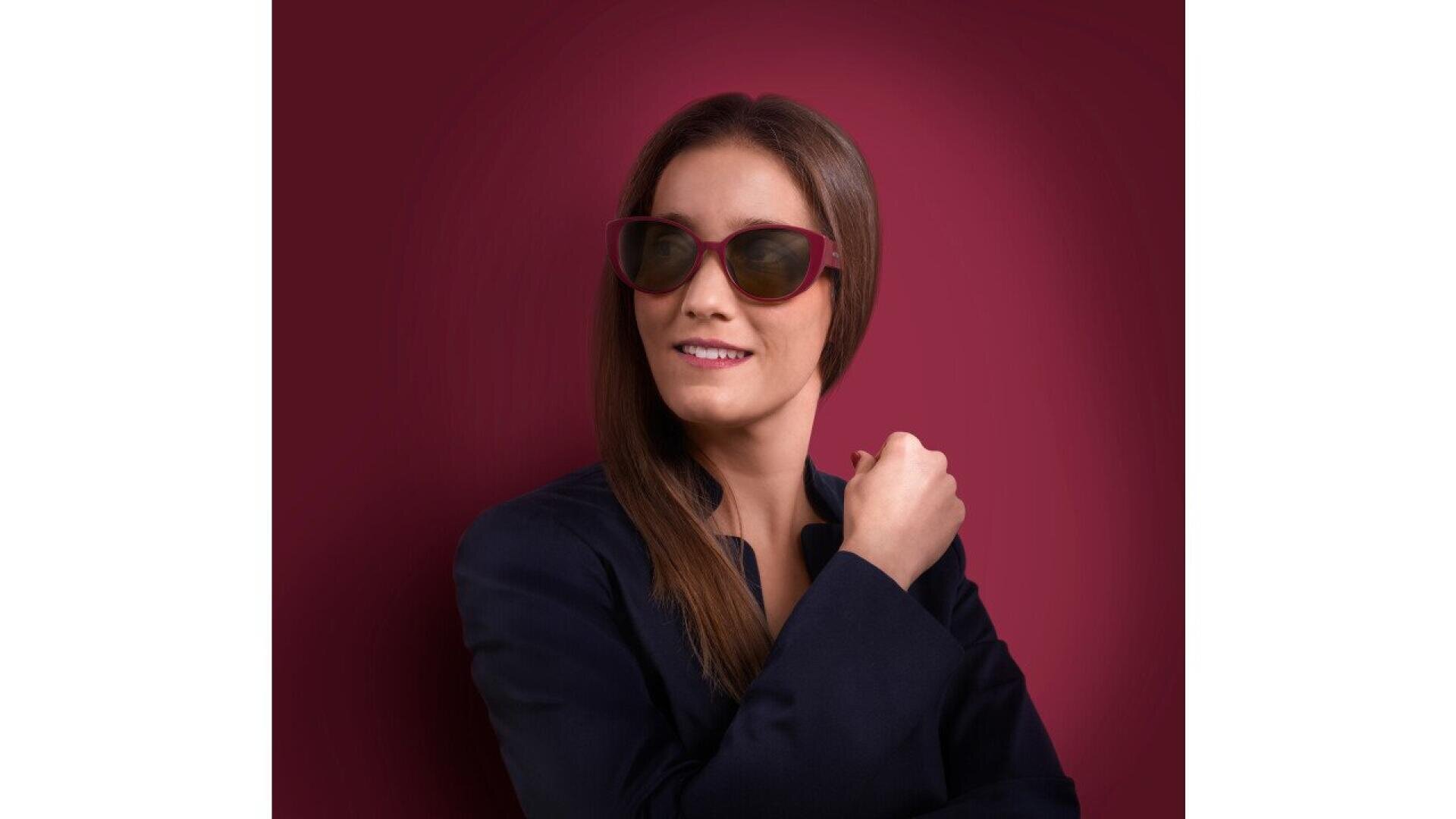A woman with long brown hair wearing red sunglasses and a dark jacket poses against a maroon background, smiling slightly and looking to the side.