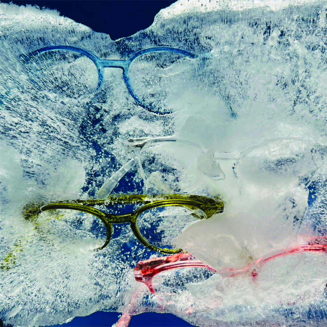 Four pairs of colorful eyeglasses—blue, clear, green, and pink—are frozen in a block of ice against a dark background. The glasses are partially visible through the icy surface.