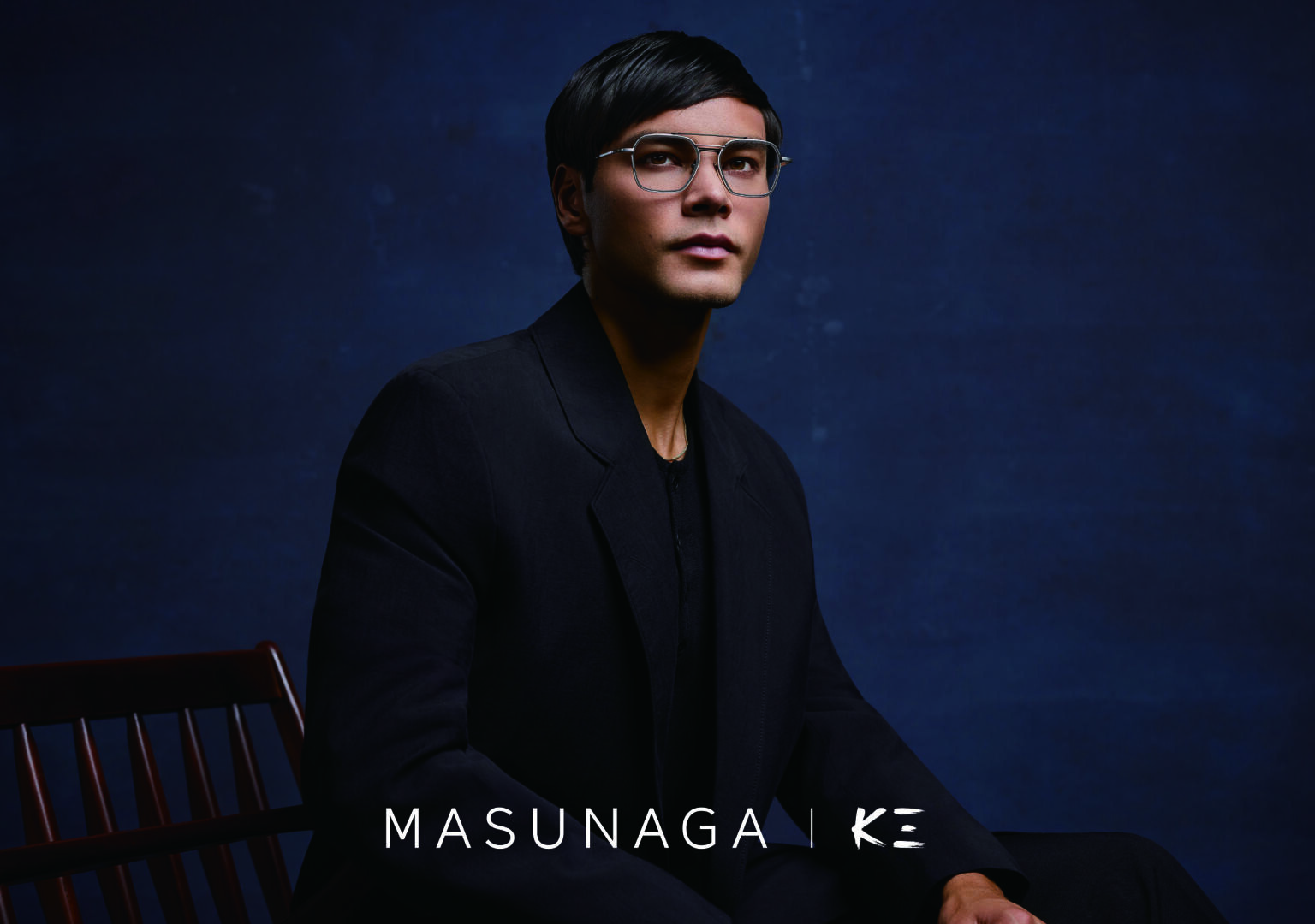 Eine Person mit kurzen dunklen Haaren, die eine Brille und einen schwarzen Anzug trägt, sitzt auf einem Holzstuhl vor einem dunkelblauen Hintergrund. Der Text MASUNAGA | KE erscheint am unteren Rand des Bildes.