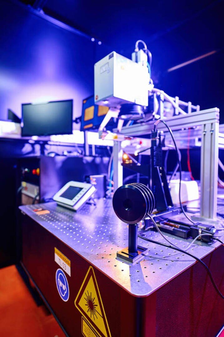 High-tech laboratory set-up with a laser warning sign, electronic devices, screens and scientific instruments on a perforated metal table, illuminated by blue and violet light.