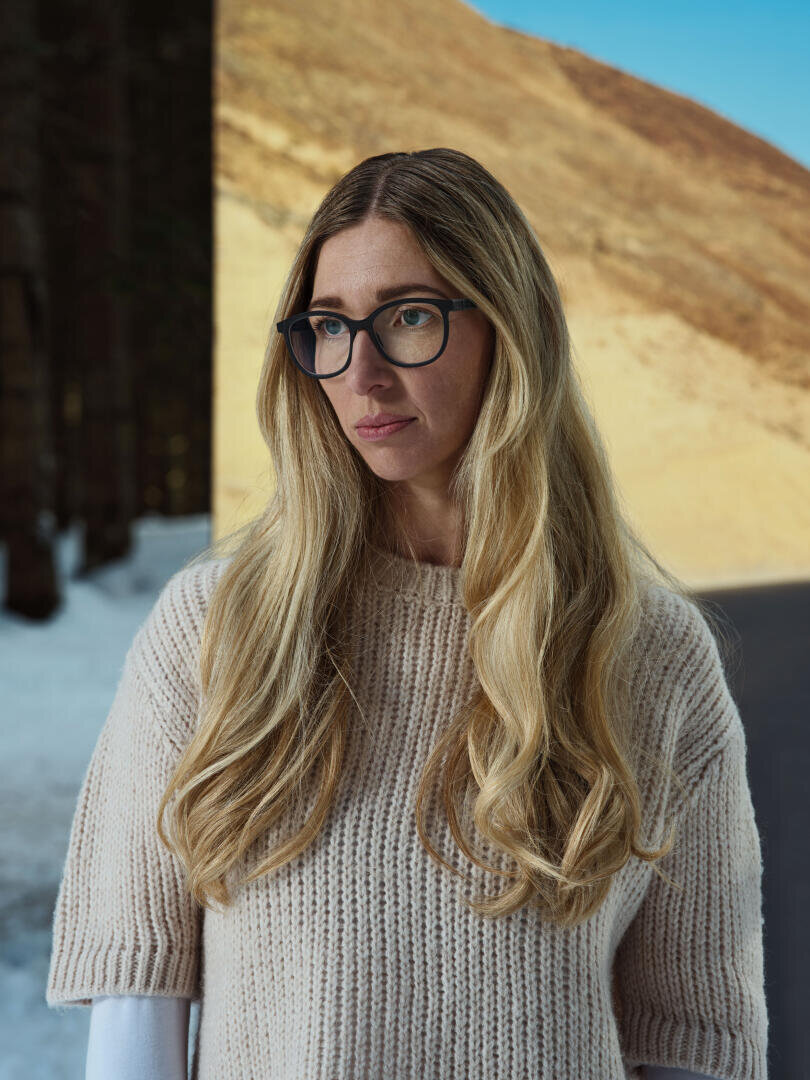 A woman with long blonde hair and glasses is standing outside wearing a beige knitted sweater. Behind her is a high-contrast background with a sunlit hill and dark, snow-covered trees.