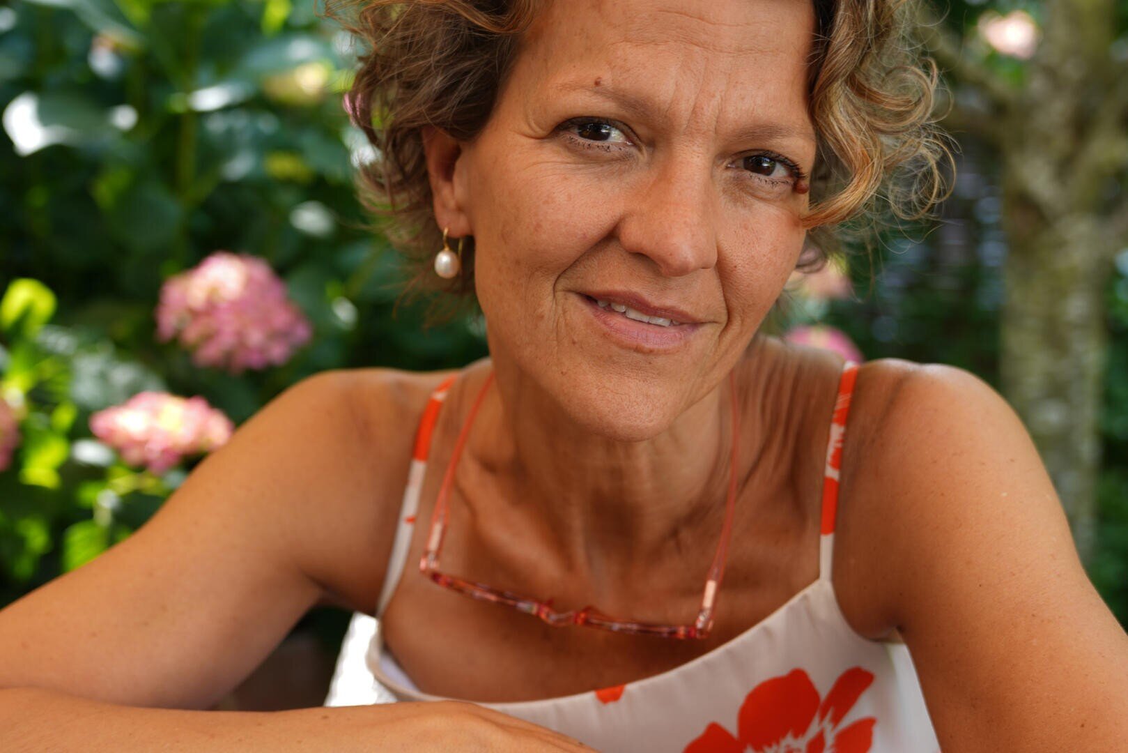 Eine Frau mittleren Alters mit lockigem Haar, die ein geblümtes Kleid und Perlenohrringe trägt, lächelt sanft, während sie im Freien vor blühenden rosa Blumen und grünem Laub sitzt.