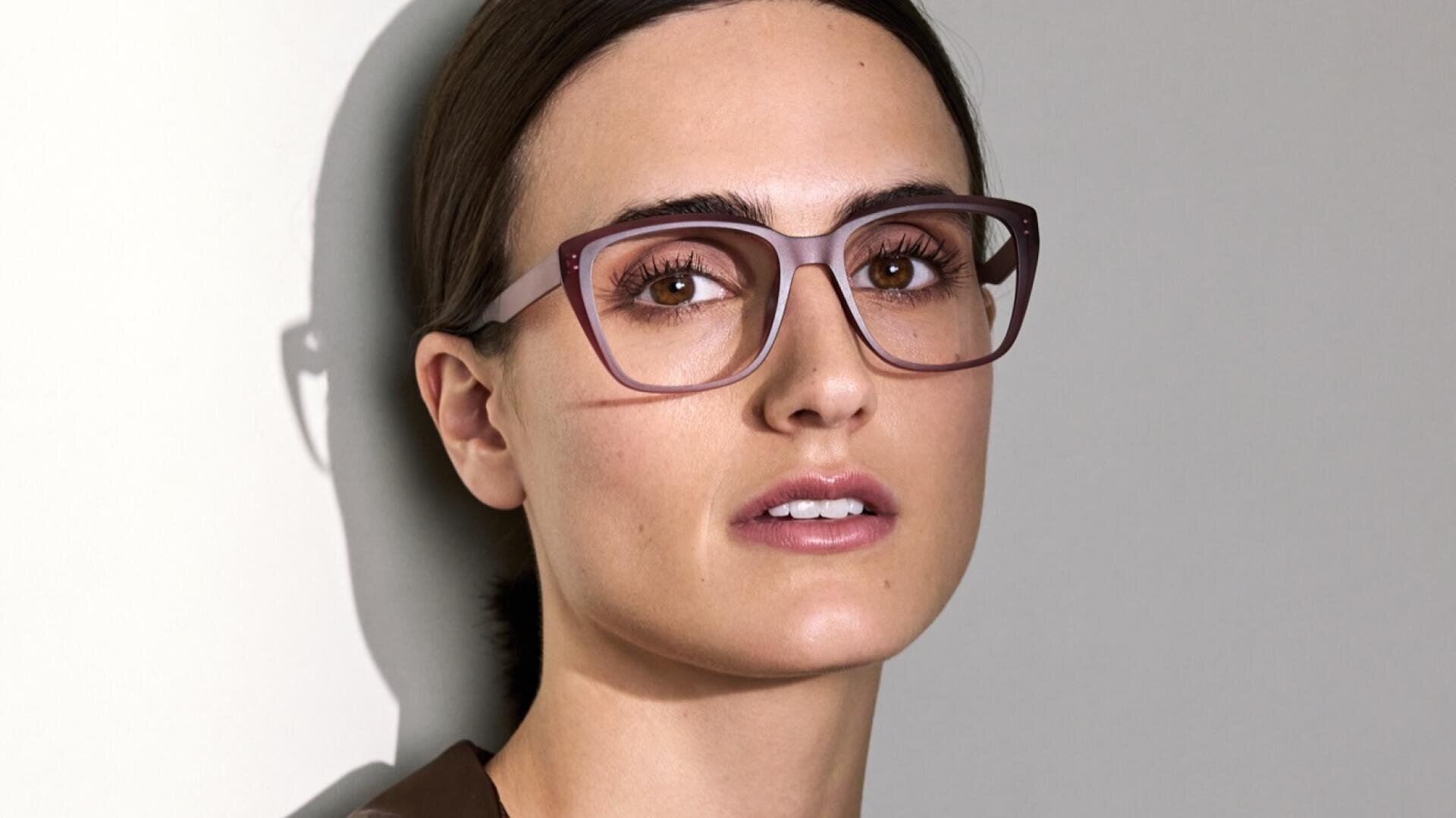 Eine Person mit heller Haut und dunklem, nach hinten gezogenem Haar trägt eine große, hellrosa Brille und blickt mit neutralem Gesichtsausdruck direkt in die Kamera vor einem hellen Hintergrund.