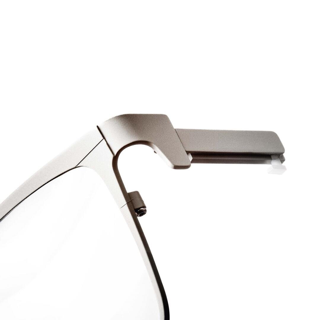 Nahaufnahme des linken Scharniers einer modernen, silbernen Brille mit rechteckigem Rahmen. Das Scharnier scheint ein einzigartiges, mechanisches Design mit einem kleinen, vorstehenden weißen Teil am Ende des Bügels zu haben.