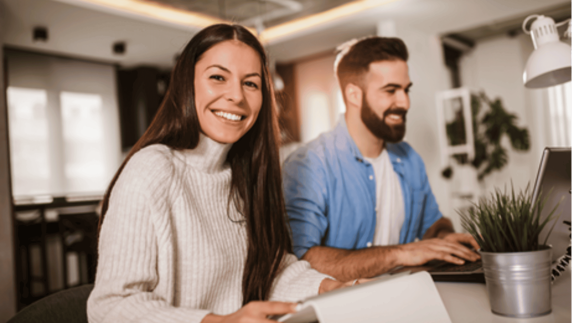 Eine Frau in einem weißen Pullover lächelt in die Kamera, während sie an einem Schreibtisch mit einem Notebook sitzt. Neben ihr arbeitet ein Mann in einem blauen Hemd an einem Laptop. Sie befinden sich in einem modernen, gut beleuchteten Büro.