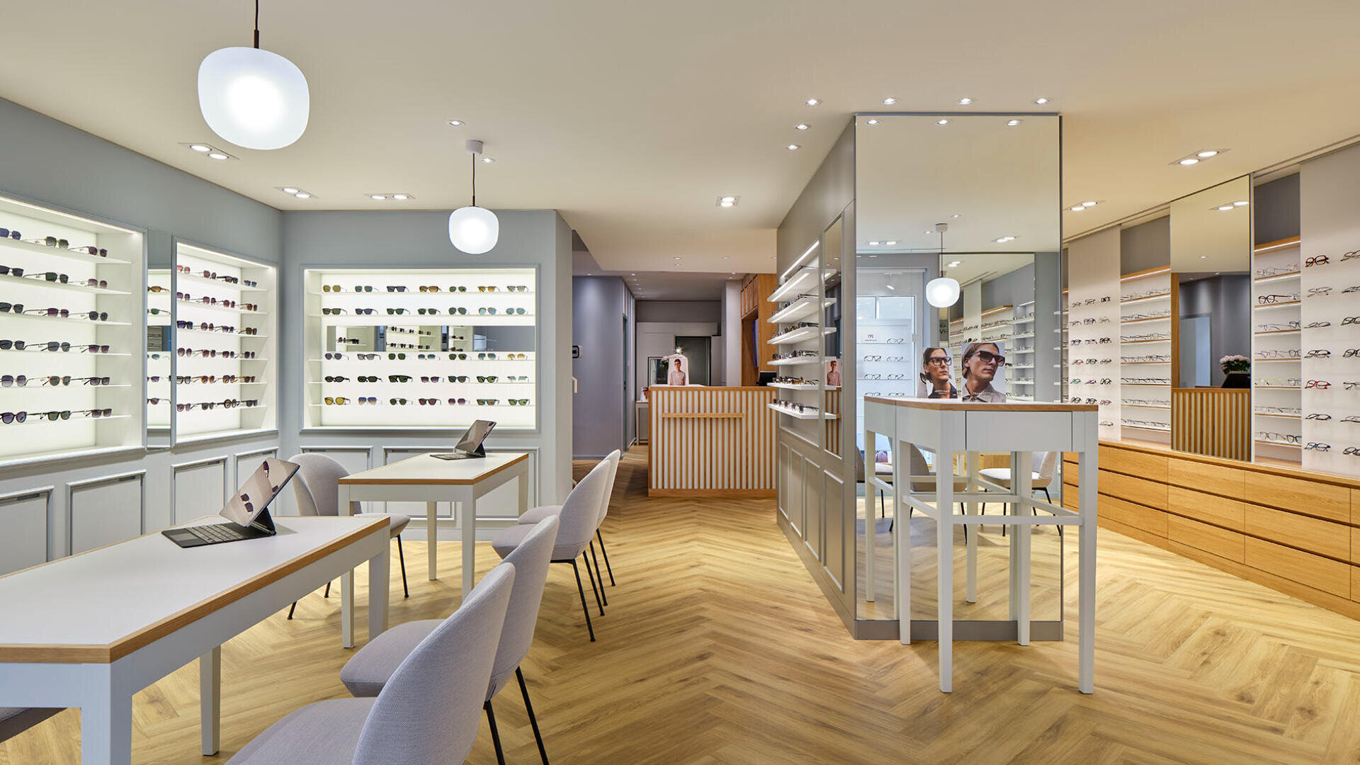 Modernes Interieur eines Brillengeschäfts mit Holzboden, mit Brillen gefüllten Regalen an den Wänden, Tischen mit Stühlen und Laptops, heller Beleuchtung und einer verspiegelten Säule in der Mitte.