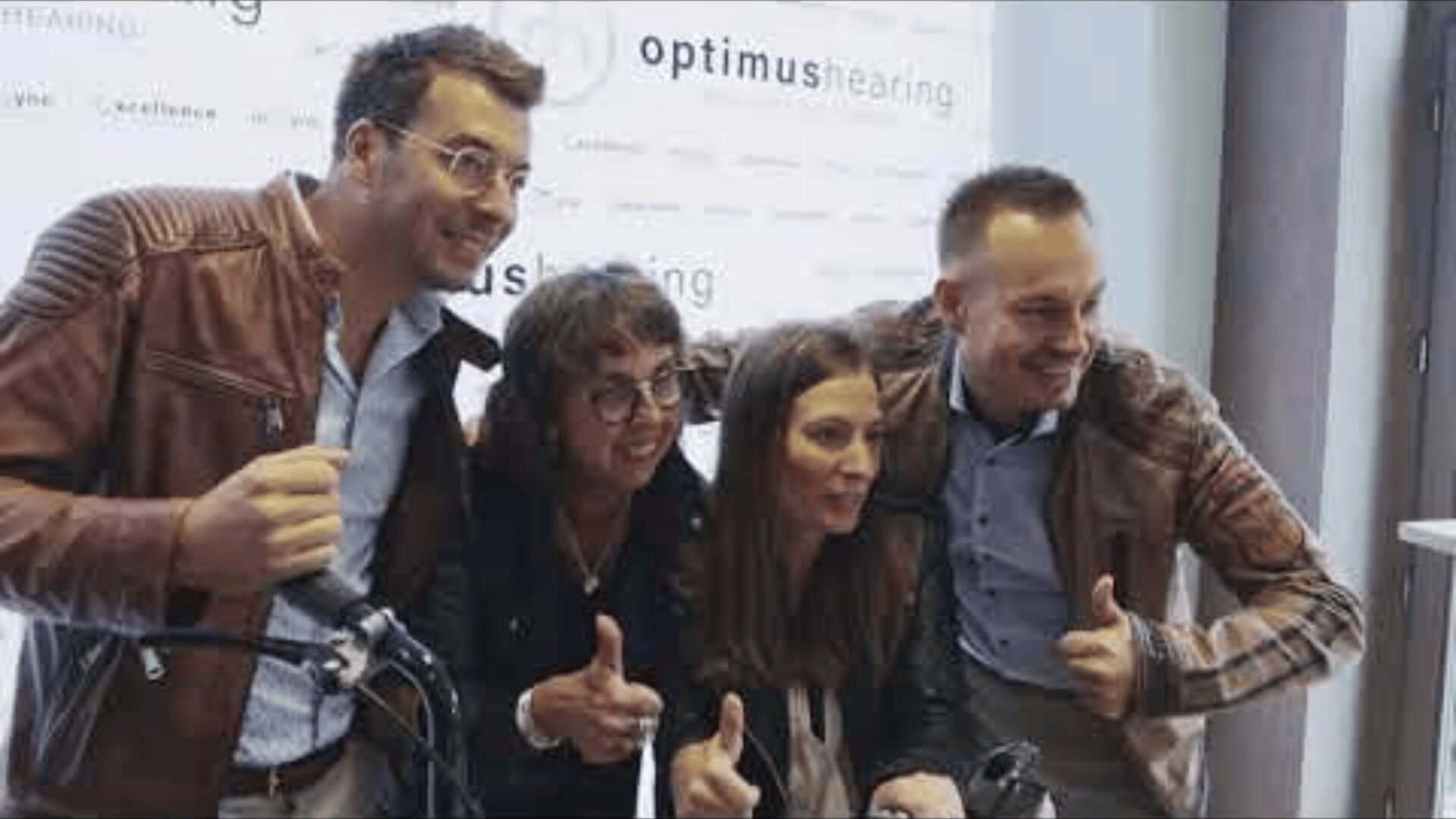 Vier Personen stehen dicht beieinander, lächeln und strecken den Daumen hoch vor einem Optimus Hearing-Banner. Sie scheinen zu feiern oder für ein Gruppenfoto bei einer Veranstaltung zu posieren.