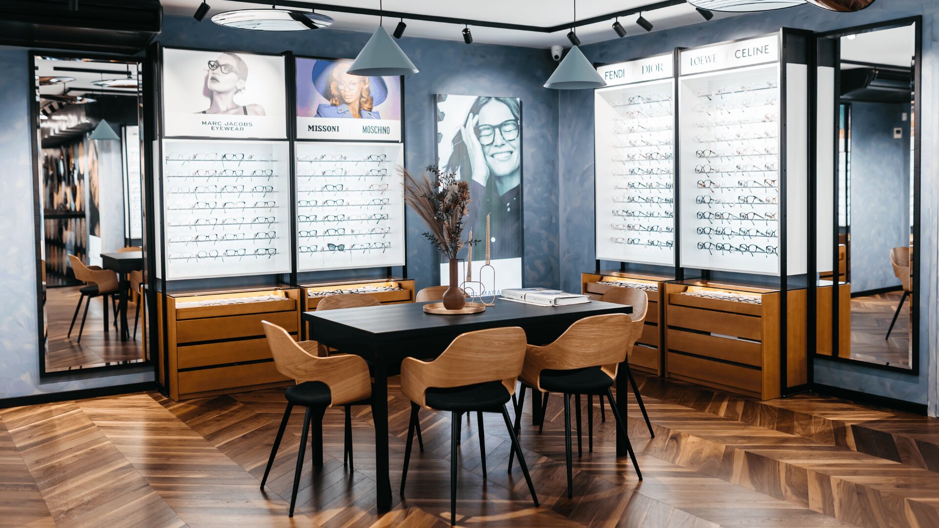 Ein modernes Brillengeschäft mit Holzboden, einem schwarzen Tisch mit Stühlen, Vitrinen mit Brillengestellen und großen Fotos von Brillenträgern an den Wänden.