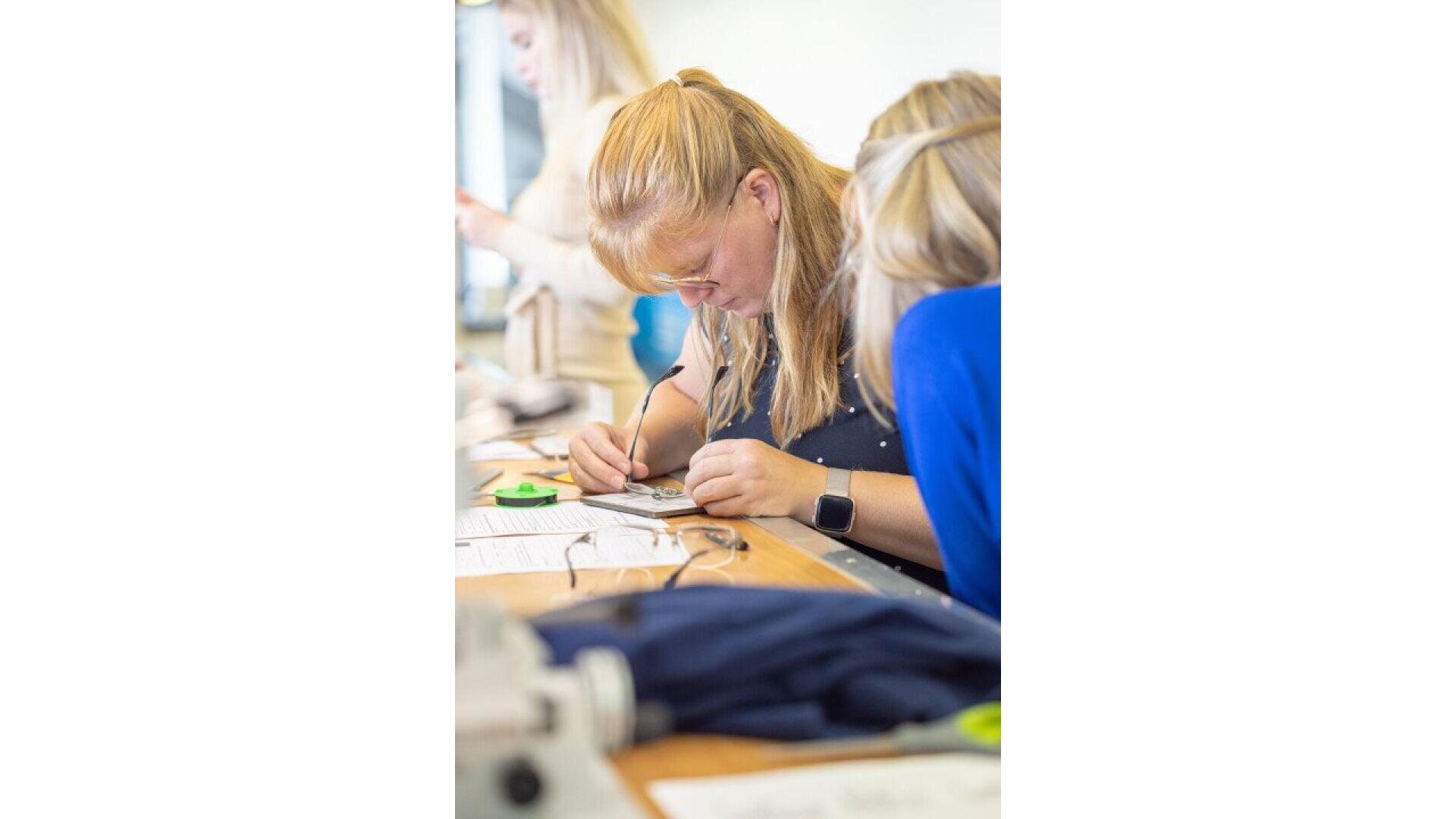 A woman with blonde hair and glasses is concentrating while working on a project at a table, using a tool and closely examining her work. Other people are also working in the background.