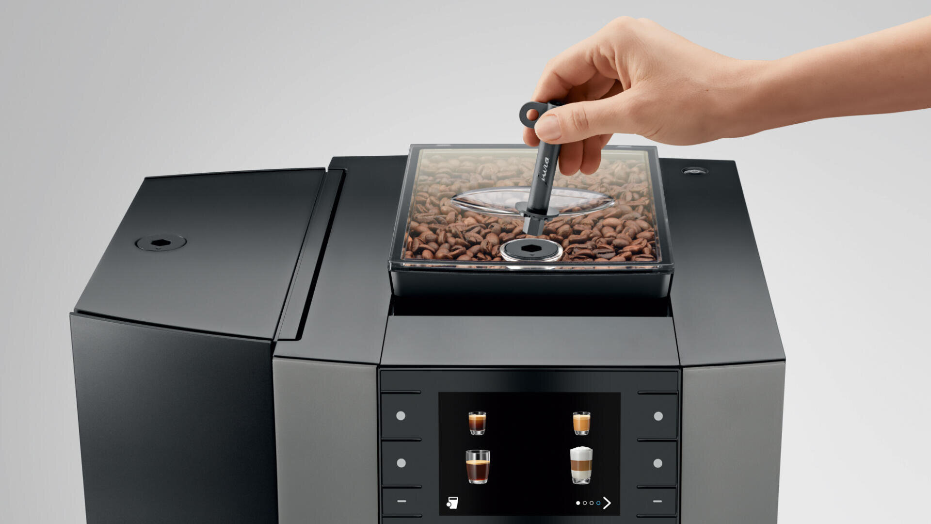 Eine Hand steckt einen Messlöffel in ein transparentes Fach mit Kaffeebohnen auf einem modernen Kaffeevollautomaten mit einer digitalen Anzeige der Getränkeoptionen.
