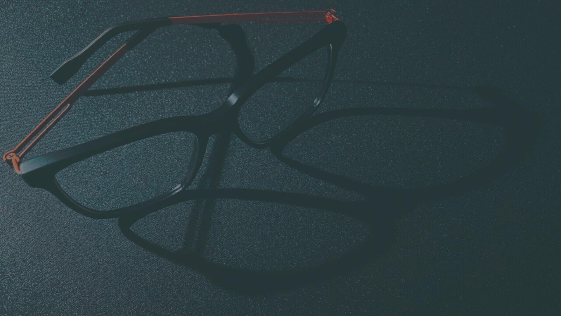 A pair of black eyeglasses with thin red arms casts a double shadow on a dark, textured surface under dim lighting.