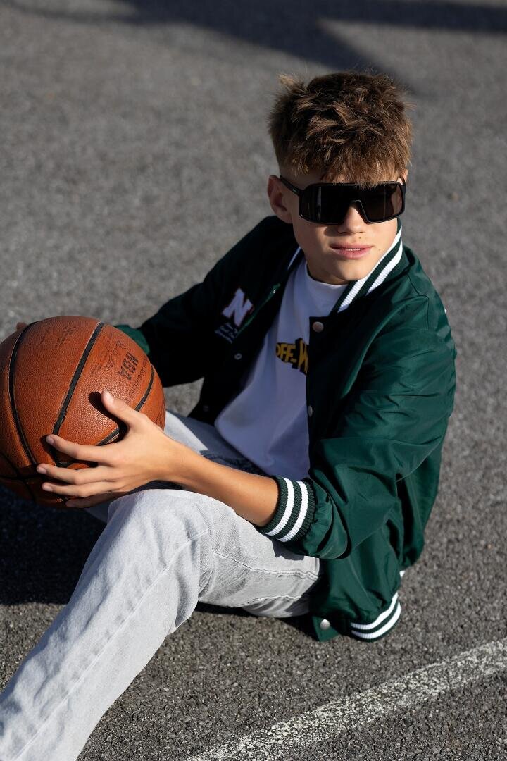 Ein Junge mit großer Sonnenbrille, grüner Jacke und hellen Jeans sitzt auf einem Basketballplatz im Freien, hält einen Basketball und blickt mit entspannter Miene zur Seite.