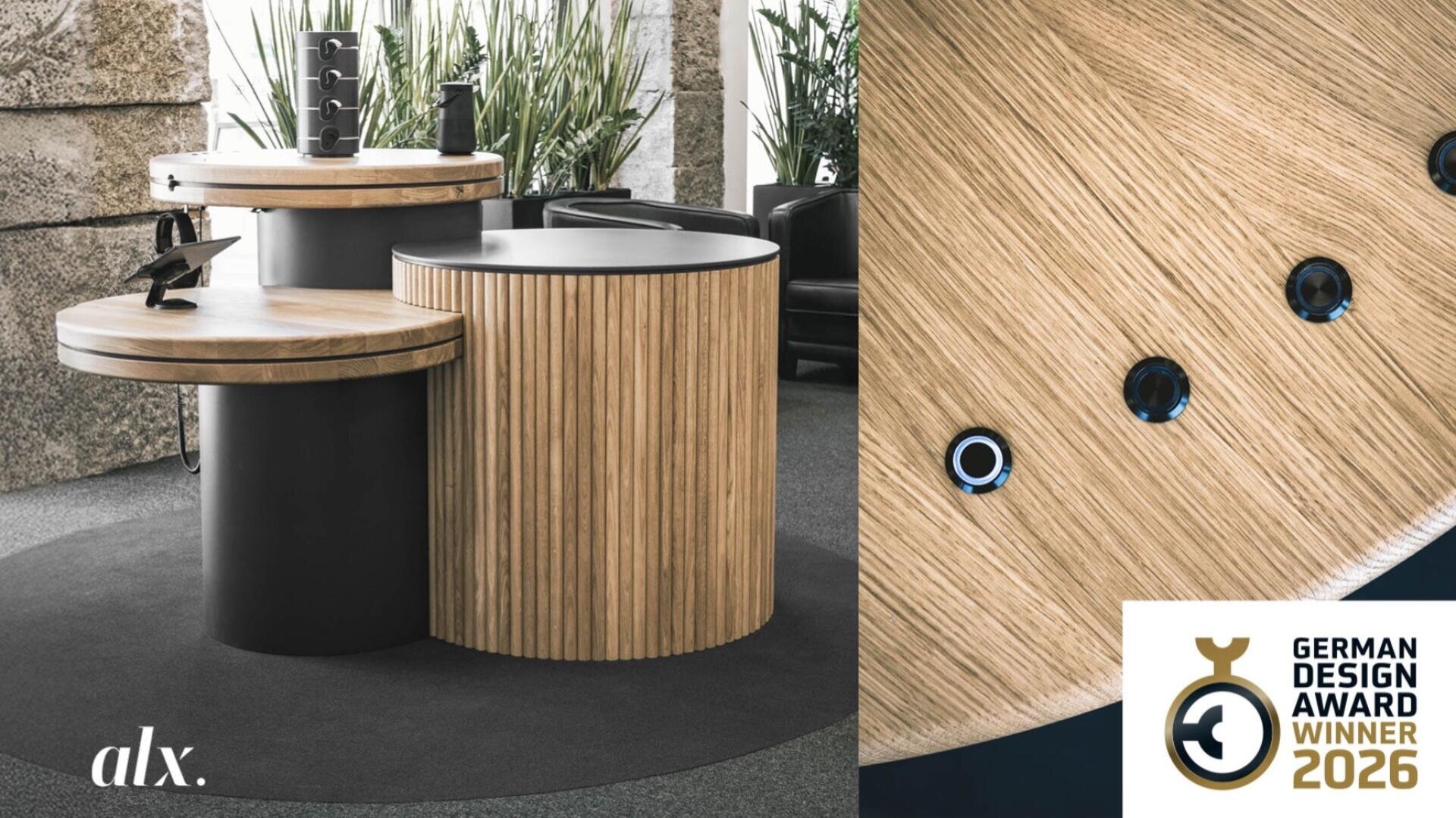 A modern, round wooden table with black accents is displayed in an interior surrounded by plants. A plaque of the German Design Award Winner 2026 can be seen in the bottom right corner, alx. can be seen in the bottom left corner.