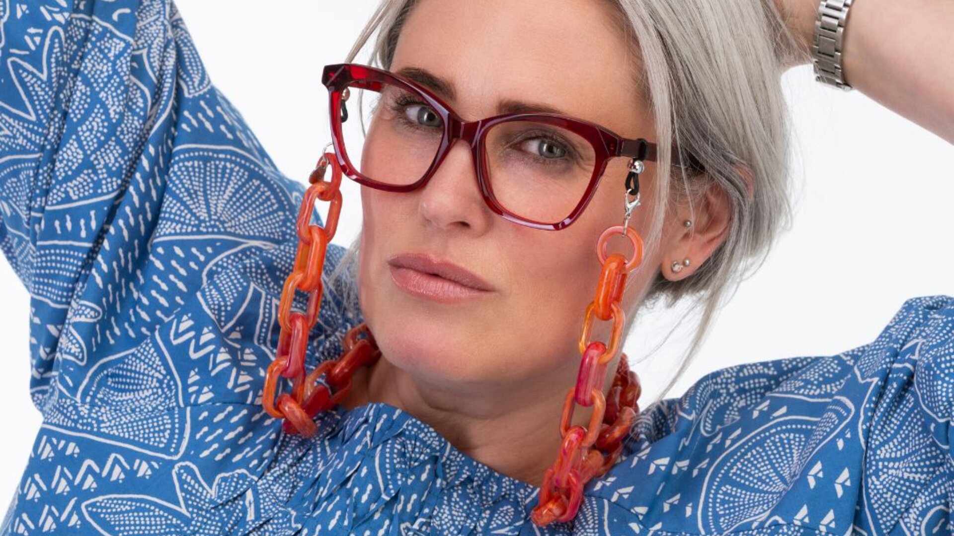 A woman with gray hair wears red glasses attached to a chunky orange chain. She is dressed in a blue patterned shirt and is posing with her hands behind her head, looking confidently at the camera.