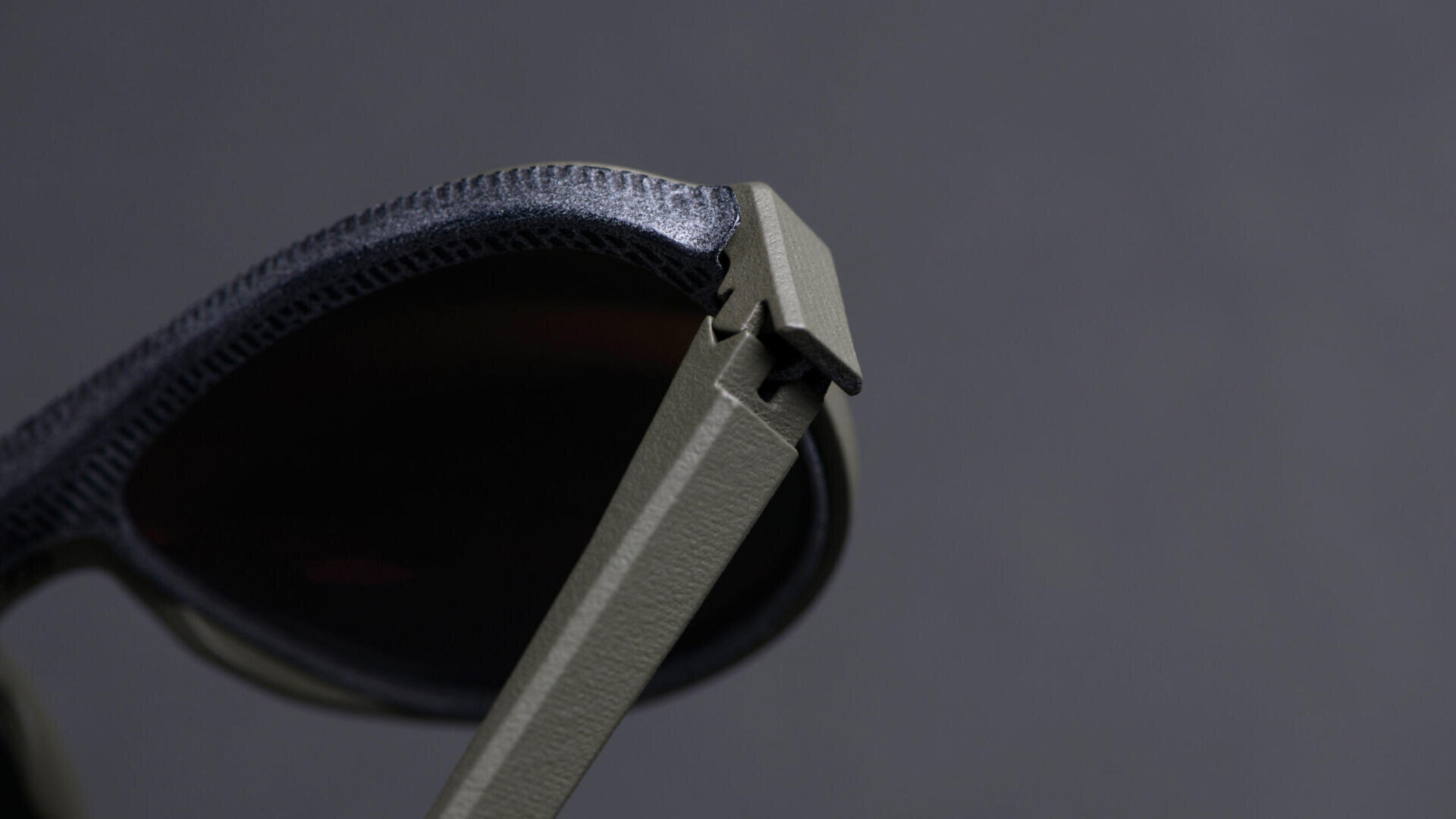 Close-up of a pair of sunglasses with dark lenses and a textured frame, focusing on the hinge where the frame and temple arm connect, against a plain gray background.