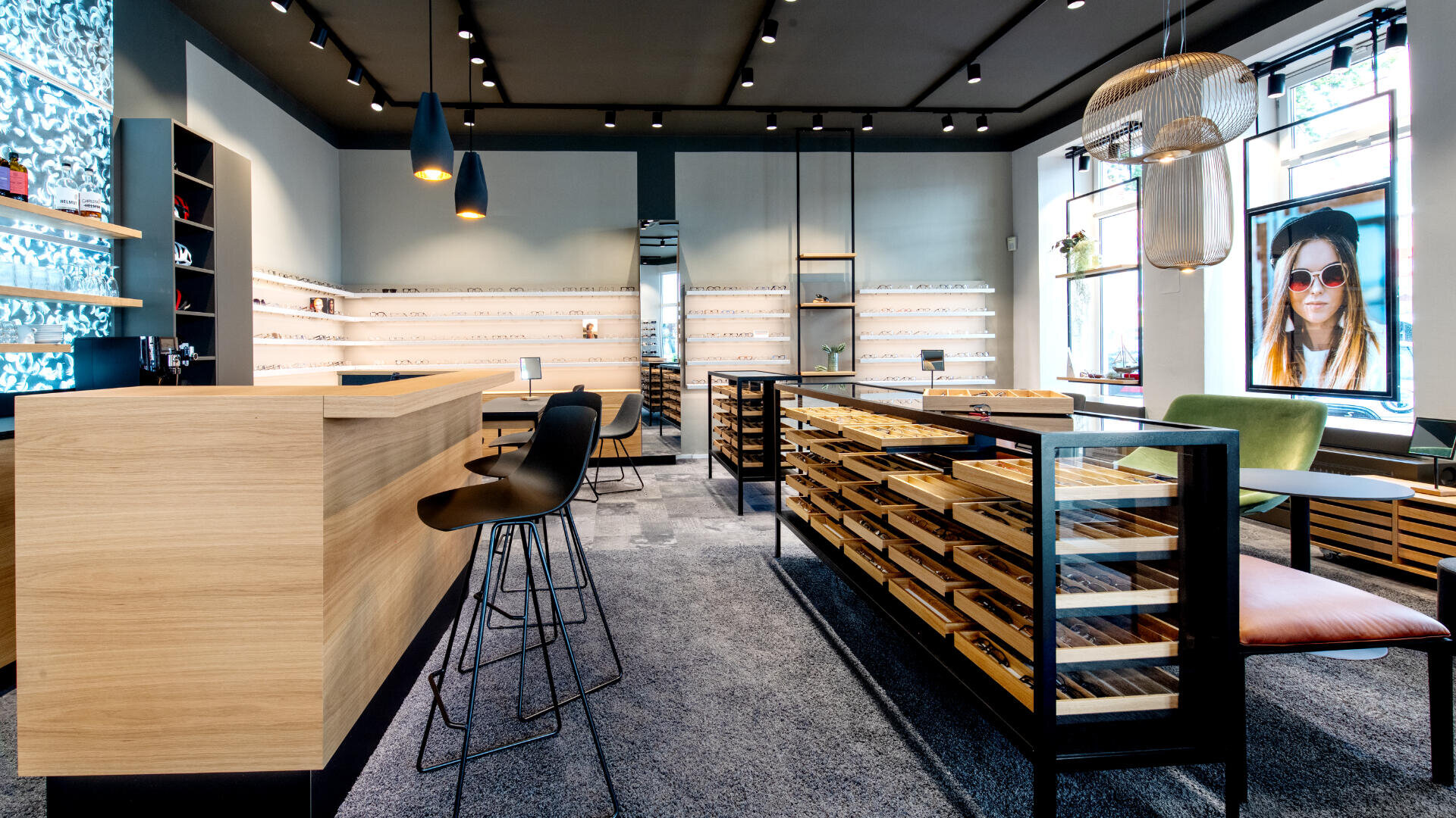 Ein modernes, gut beleuchtetes Optikergeschäft mit Holzvitrinen, in denen reihenweise Brillengestelle ausgestellt sind, eleganten schwarzen Stühlen, Hängelampen und großen Fenstern. Draußen ist durch ein Fenster eine Frau mit Sonnenbrille zu sehen.