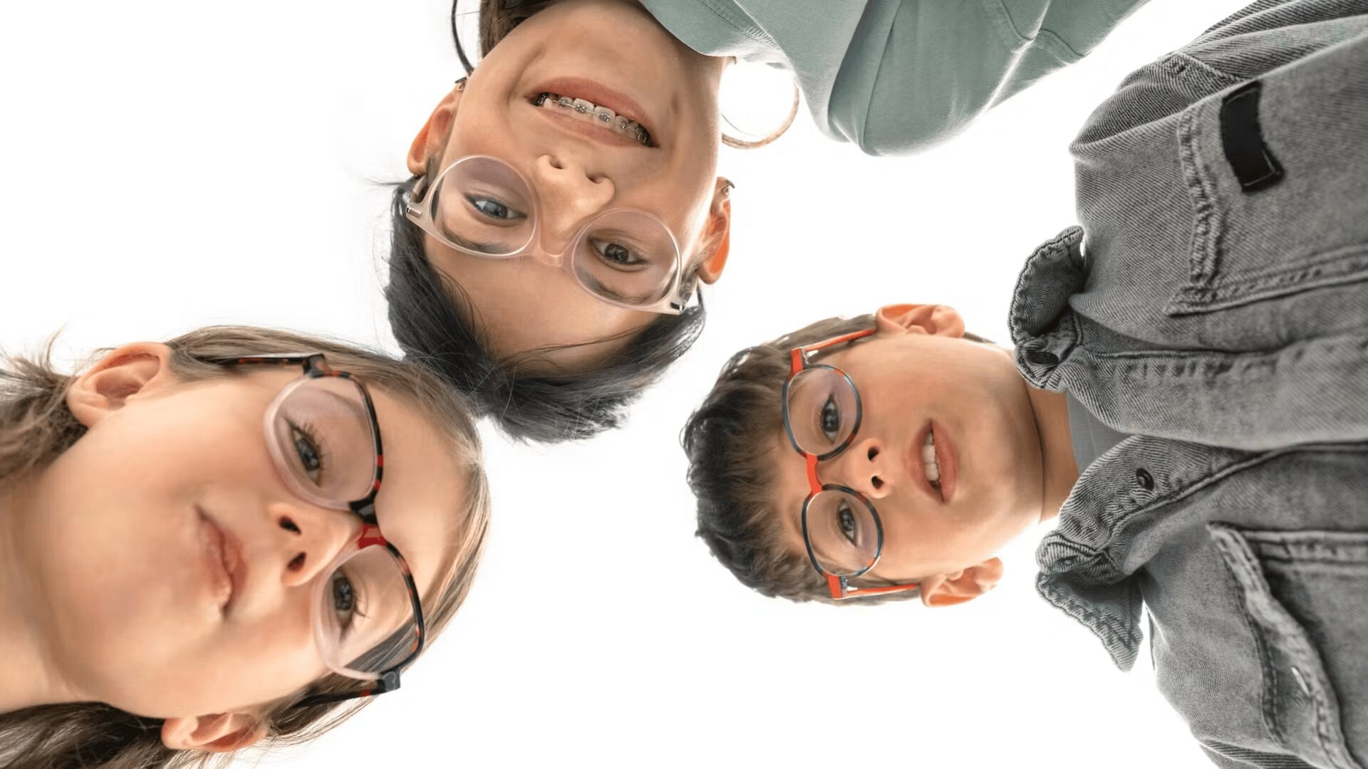 Three children wearing glasses are standing in a circle, looking down at the camera and smiling against a white background. The image is taken from a low angle, capturing their faces closely.