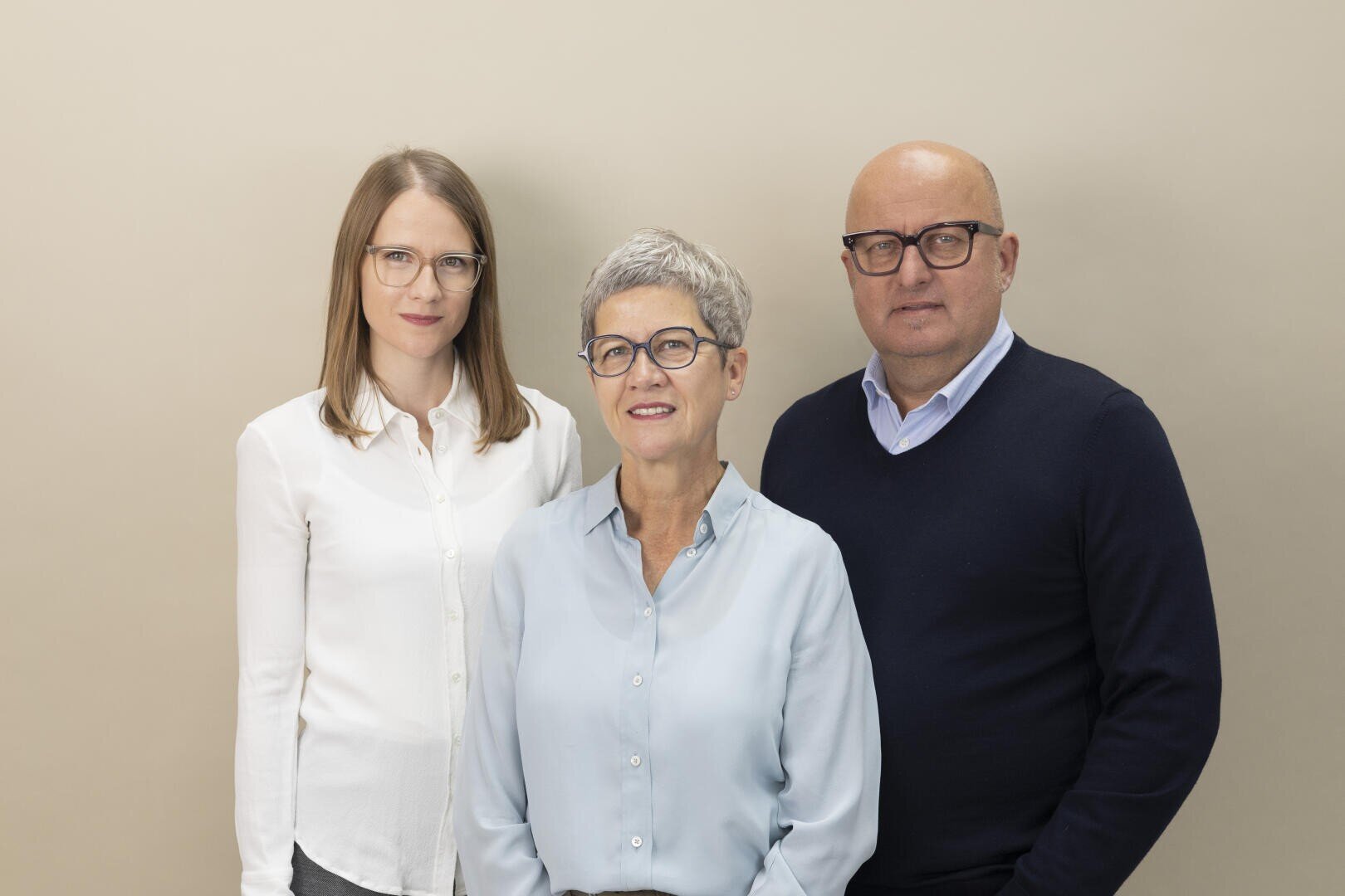 Three adults stand together in front of a plain beige background. Two women, one younger and one older with short gray hair, and an older man, all wearing glasses and casual business attire, look into the camera and smile gently.
