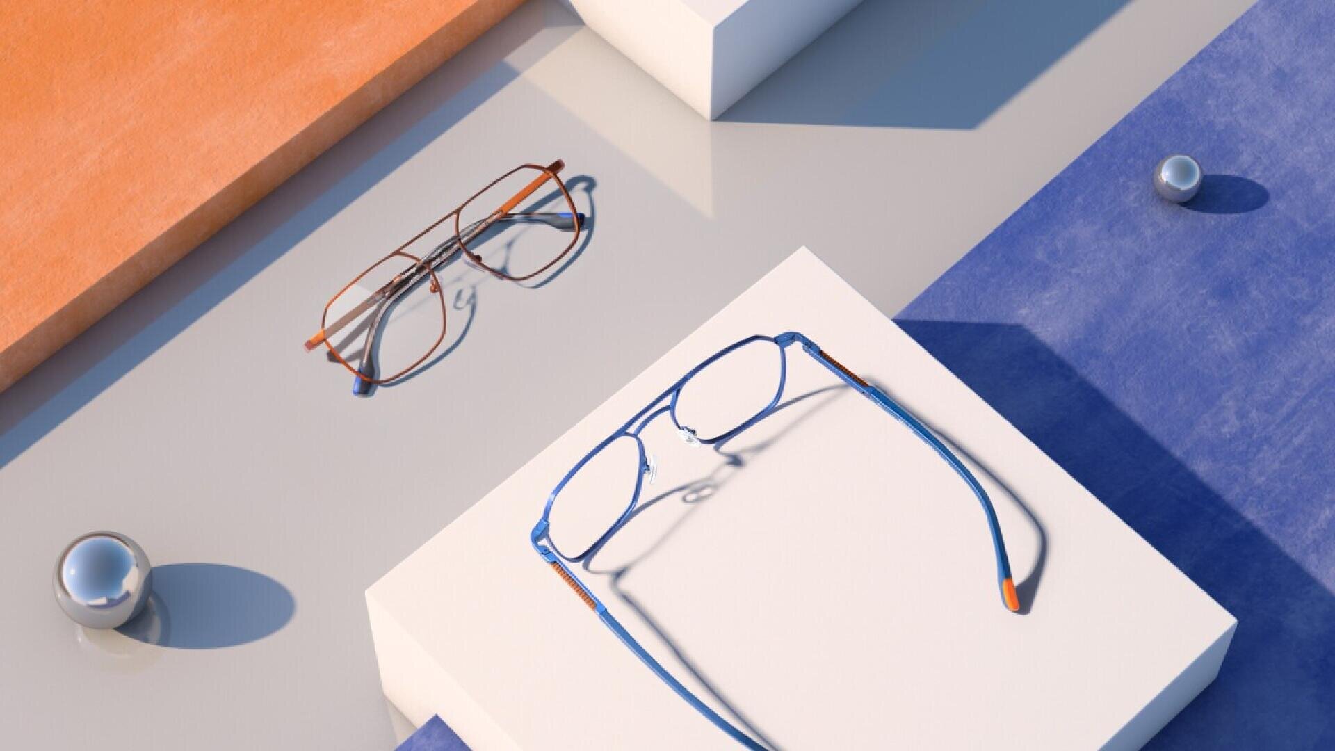 Two pairs of eyeglasses, one brown and one blue, are displayed on geometric white platforms with orange and blue textured backgrounds and two small metallic spheres nearby.