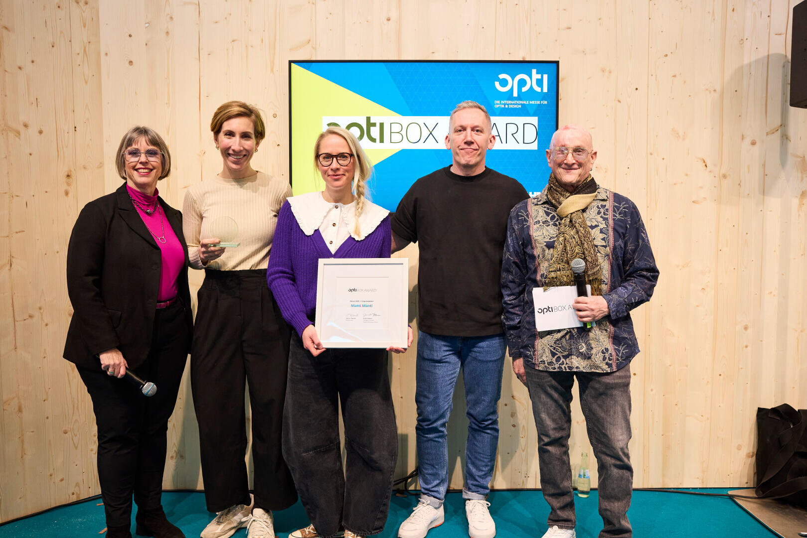 Fünf Personen stehen gemeinsam im Innenbereich vor einer Holzwand und einem Bildschirm mit der Aufschrift „opti BOX AWARD“. Eine Person in der Mitte hält eine gerahmte Urkunde, eine andere ein Mikrofon. Alle lächeln in die Kamera.