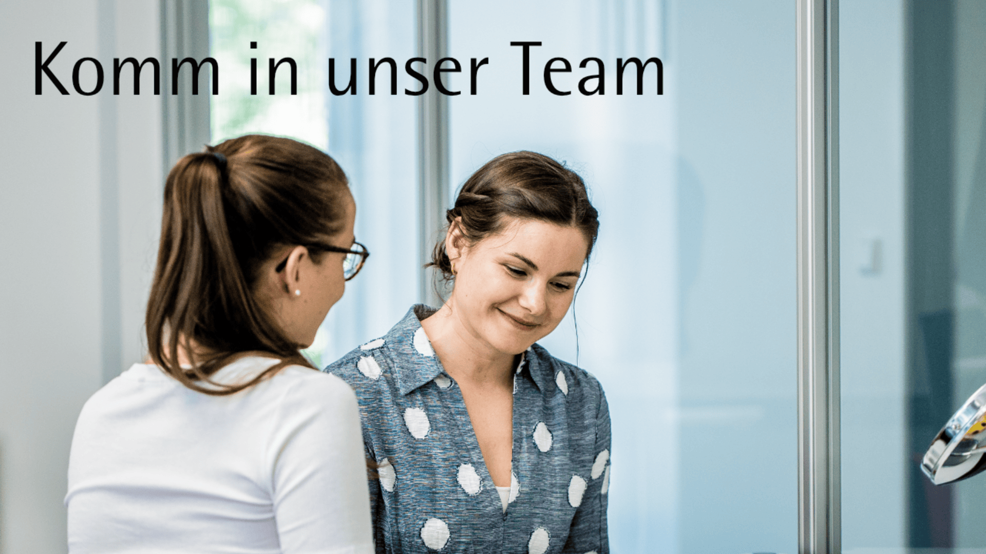 Zwei Frauen in einer Büroumgebung lächeln und unterhalten sich. Der Text "Komm in unser Team" wird oben angezeigt und lädt Menschen ein, ihrem Team beizutreten.