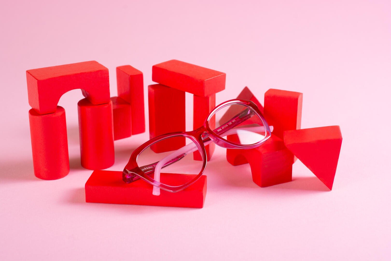 Eine rote Brille ruht auf roten geometrischen Holzblöcken, die auf einer rosa Fläche mit rosa Hintergrund angeordnet sind.