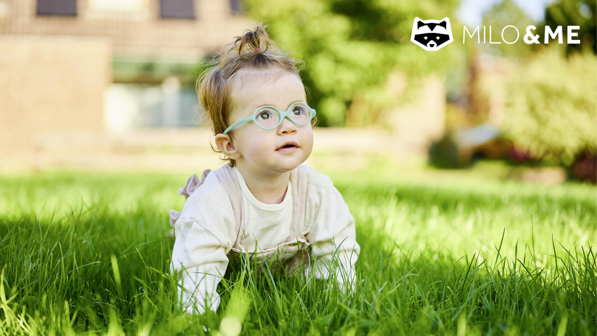 Ein Baby mit hellem Haar und Brille krabbelt im Freien auf grünem Gras. Das Baby trägt ein helles Outfit. Der Hintergrund ist unscharf, und das MILO & ME-Logo erscheint in der oberen rechten Ecke.