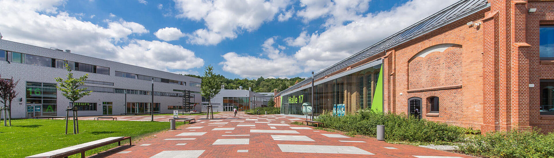 Ein weitläufiger Campus-Hof mit modernen weißen Gebäuden auf der linken und einem roten Backsteingebäude auf der rechten Seite, Bänken, Bäumen und einem blauen Himmel mit vereinzelten Wolken über dem Kopf.