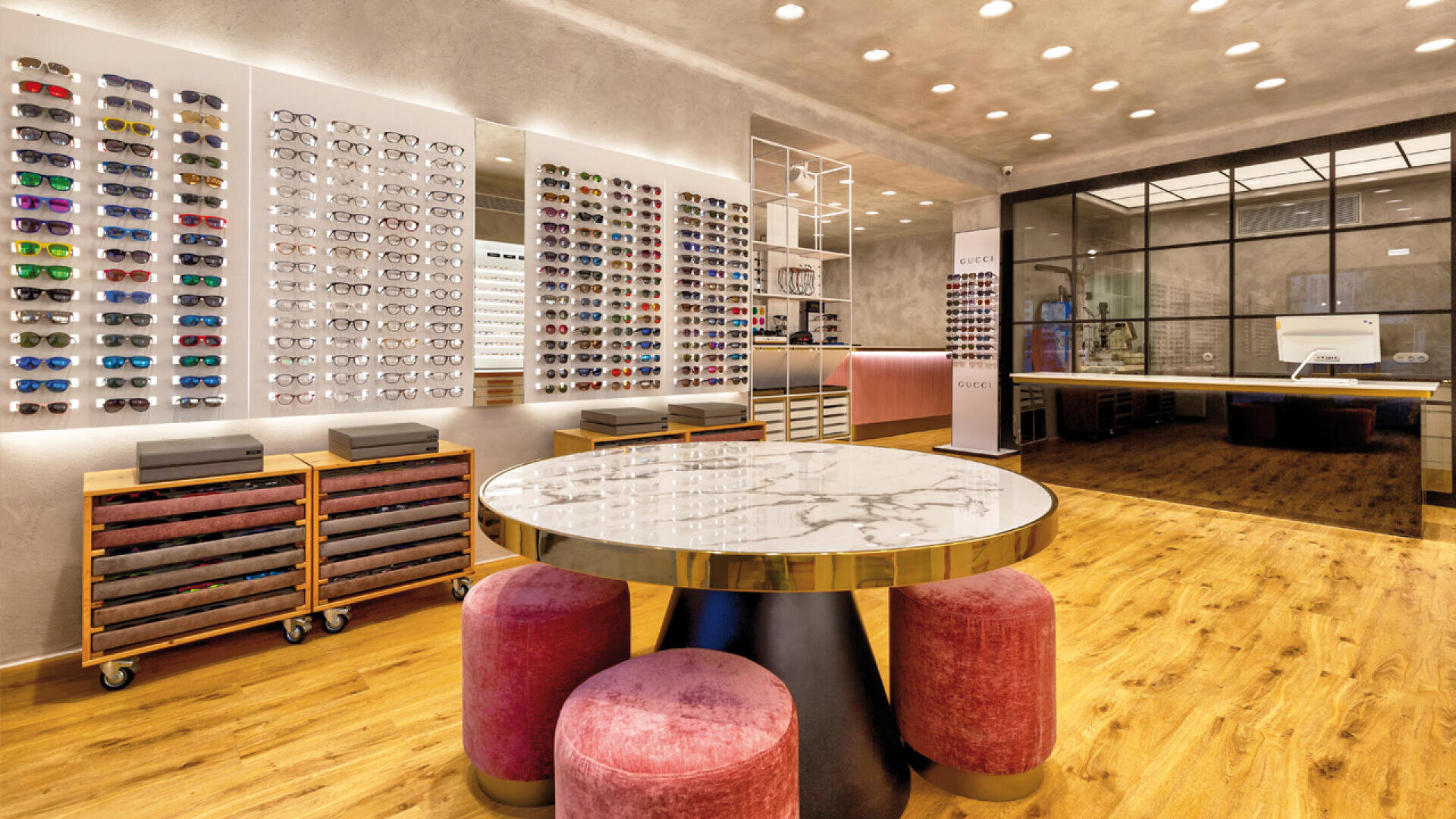 Modernes Brillengeschäft mit Holzböden, einem runden Tisch mit Marmorplatte, umgeben von rosa Hockern, Brillendisplays an der Wand, Schubladen darunter und einem geräumigen, gut beleuchteten Innenraum mit Glastrennwänden im Hintergrund.