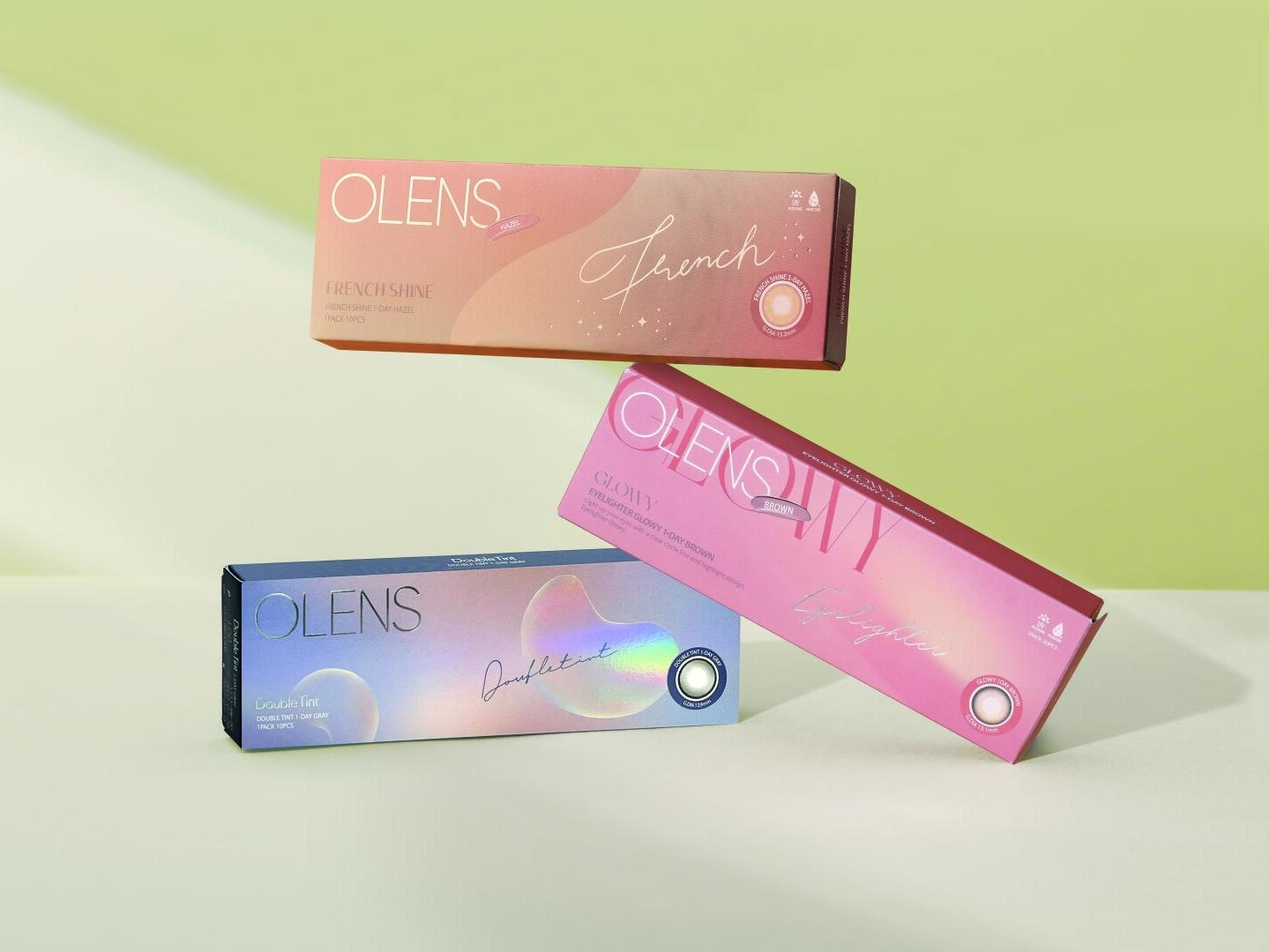 Three colorful OLENS contact lens cases can be seen against a light green background: a pink one with the inscription French Shine, a pink and coral one with the inscription Glowy and a blue and purple one with the inscription Double Tint.