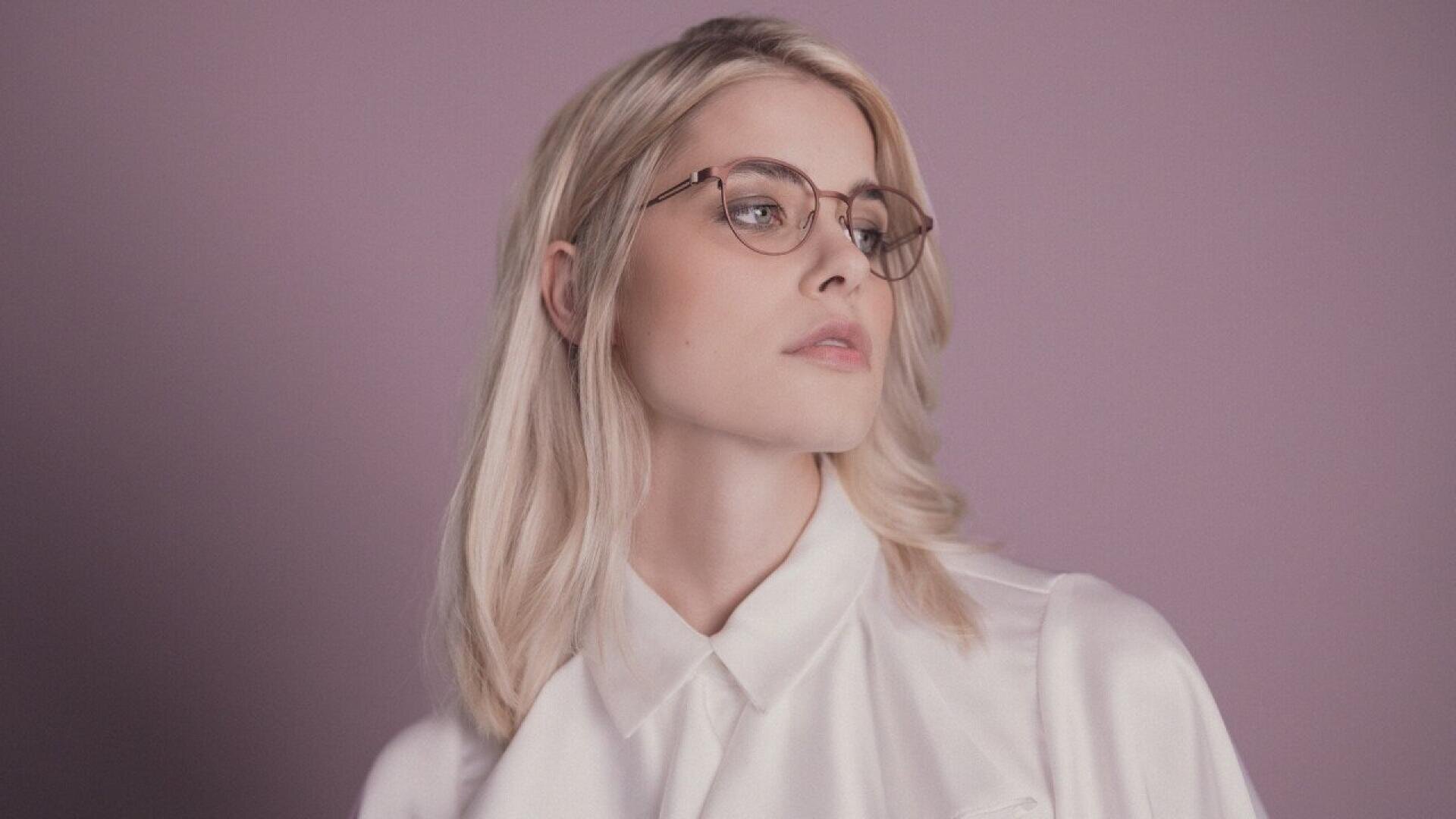 A woman with long blonde hair and glasses looks to the side. She is wearing a white blouse and is posed against a muted purple background.