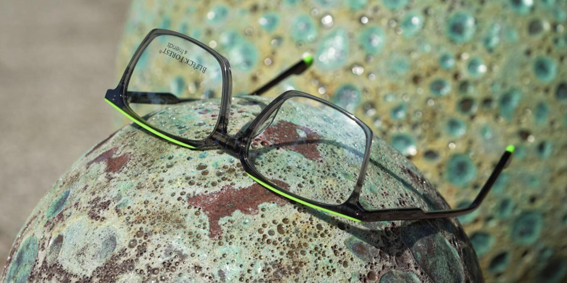 A pair of eyeglasses with black and green frames rests on a textured, greenish, weathered stone sphere. Another similar sphere appears in the blurred background.