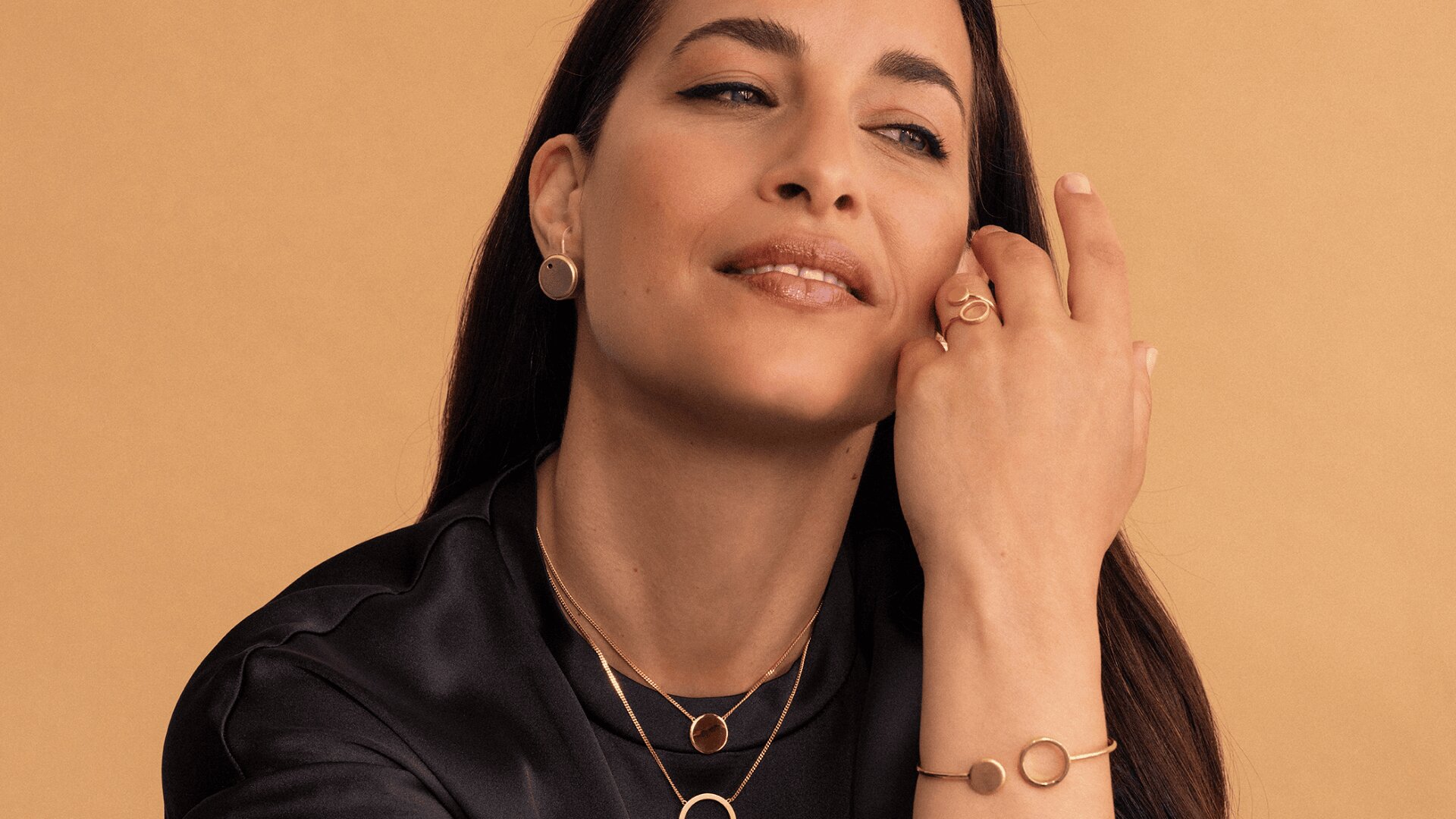 Eine Frau mit langem, dunklem Haar posiert vor einem beigen Hintergrund. Sie trägt Goldschmuck, darunter Halsketten, Ohrringe, Ringe und ein Armband mit kreisförmigen Mustern. Sie trägt ein schwarzes Oberteil und hat einen sanften, entspannten Gesichtsausdruck.