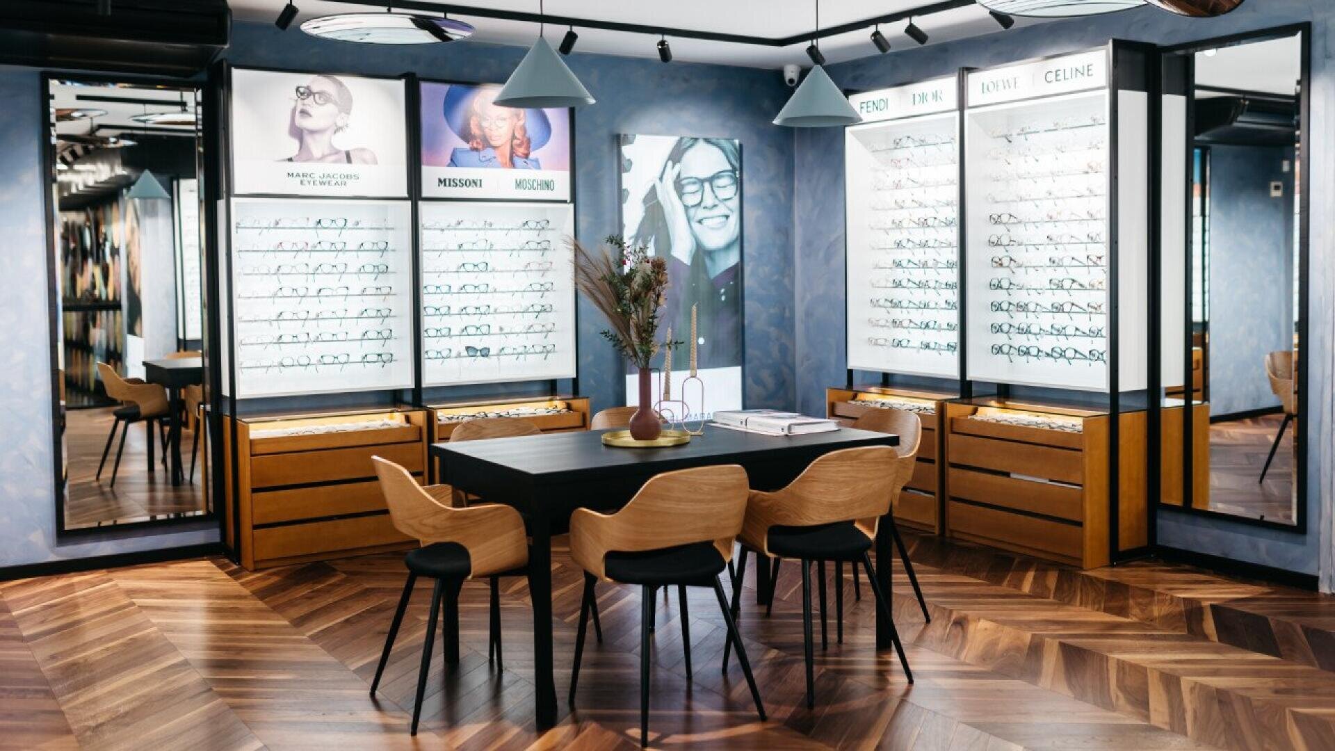 A modern optician’s shop with display cases of eyeglasses, a central black table with four wooden chairs, and decorative plants. Large photos and brand logos are on the wall behind the glasses displays.