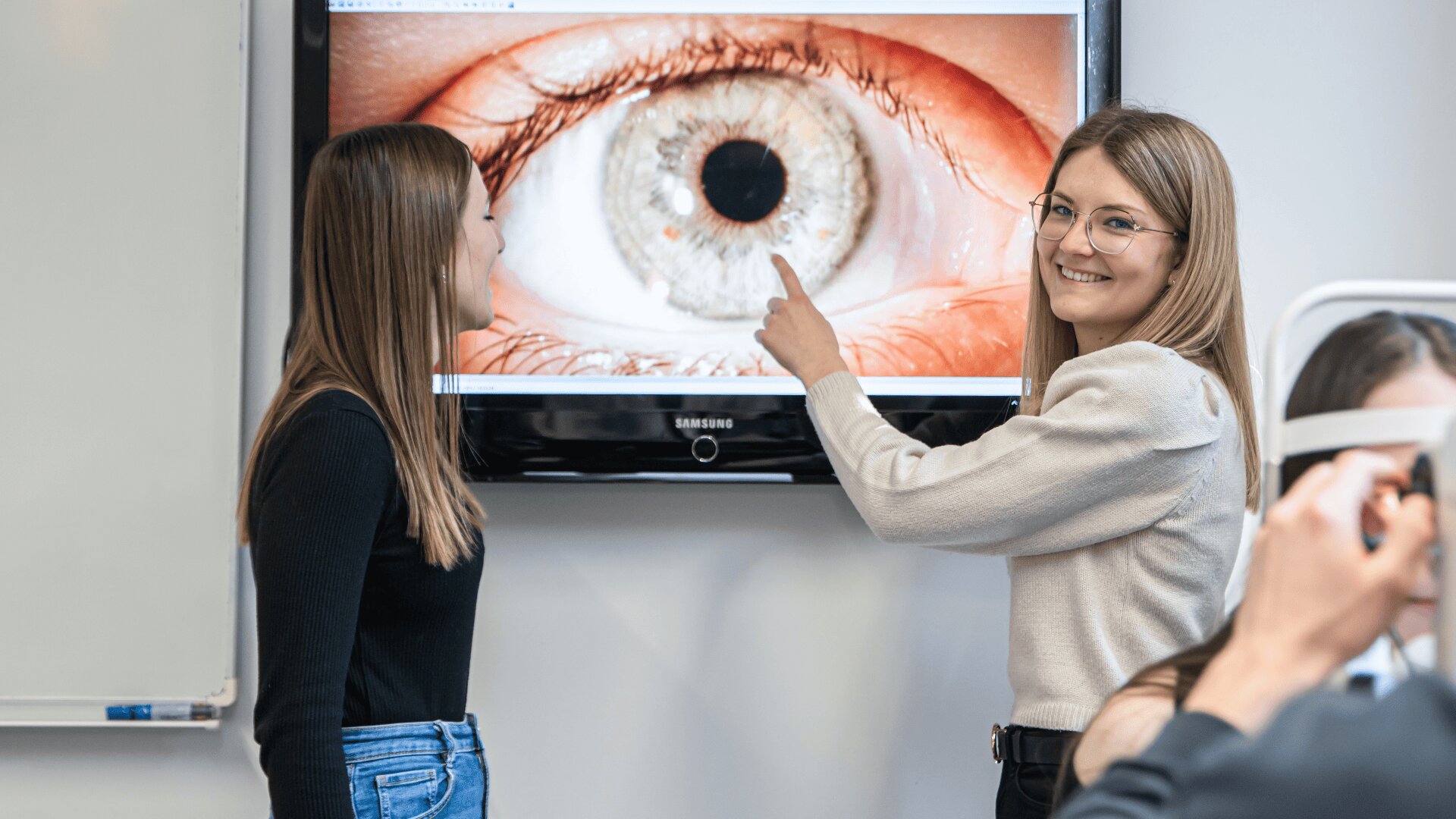 Zwei Frauen stehen vor einem großen Bildschirm, auf dem eine Nahaufnahme eines Auges zu sehen ist. Eine Frau zeigt auf den Bildschirm und lächelt, während eine andere Frau im Vordergrund sitzt und eine ophthalmologische Ausrüstung benutzt.