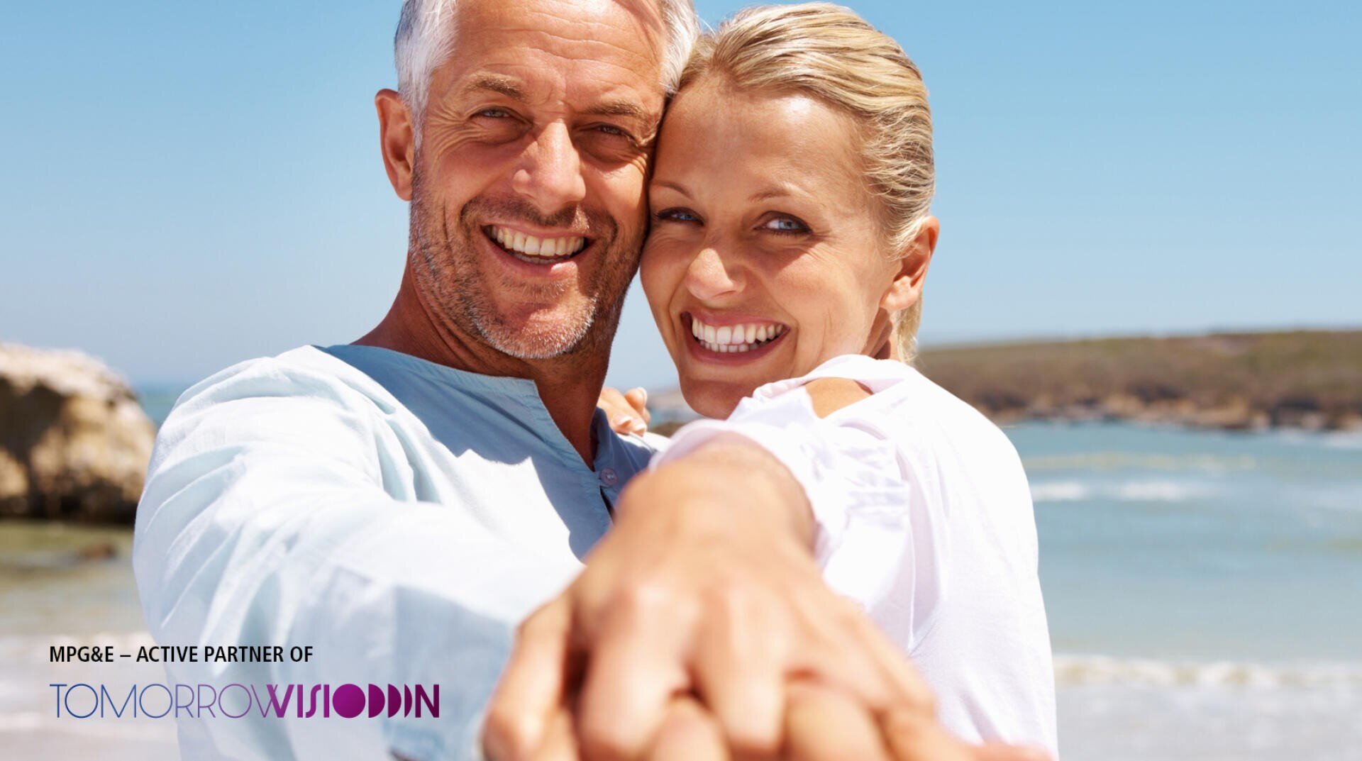 Ein lächelndes Paar stellt sich für ein Selfie an einem sonnigen Strand auf, hält sich an den Händen und schaut in die Kamera. Im Hintergrund sind das Meer und der Sandstrand zu sehen. Logos und Text erscheinen in der unteren linken Ecke.