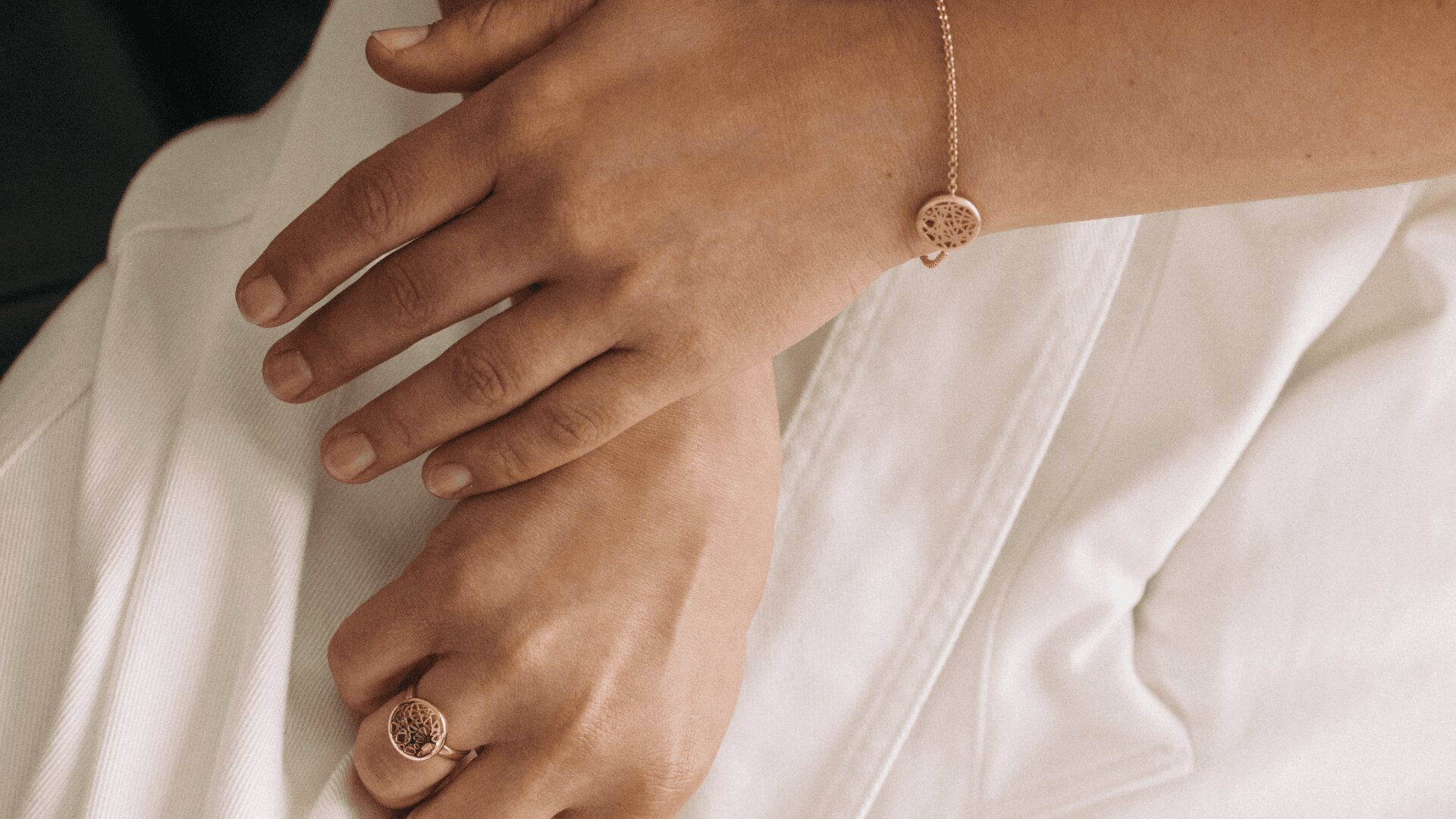 Eine Nahaufnahme zweier Hände: Eine trägt ein zartes Armband, die andere einen passenden Ring, beide mit runden, filigranen Mustern. Die Person trägt ein weißes Gewand.