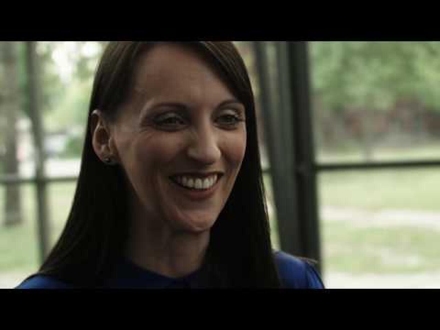 A woman with straight dark hair, wearing a blue top, smiles in an interior space in front of large windows with a blurred green background outside.
