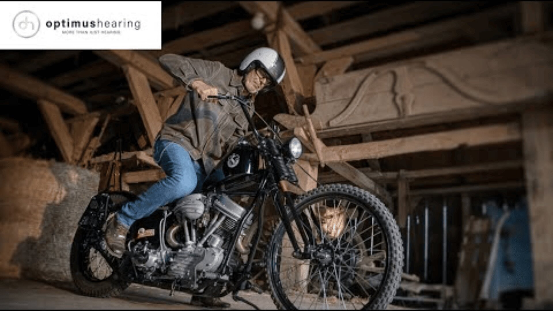 Ein Mann mit Helm und Brille startet in einer Holzscheune ein Oldtimer-Motorrad. Das Logo von optimushearing erscheint in der oberen linken Ecke.