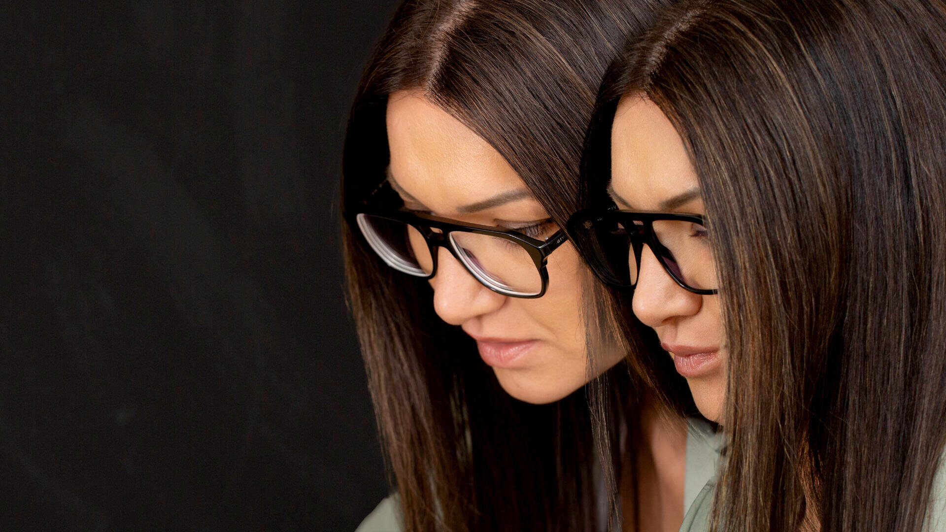 A woman with straight brown hair and glasses is facing downward, appearing twice side by side, as if reflected or duplicated, against a dark background.