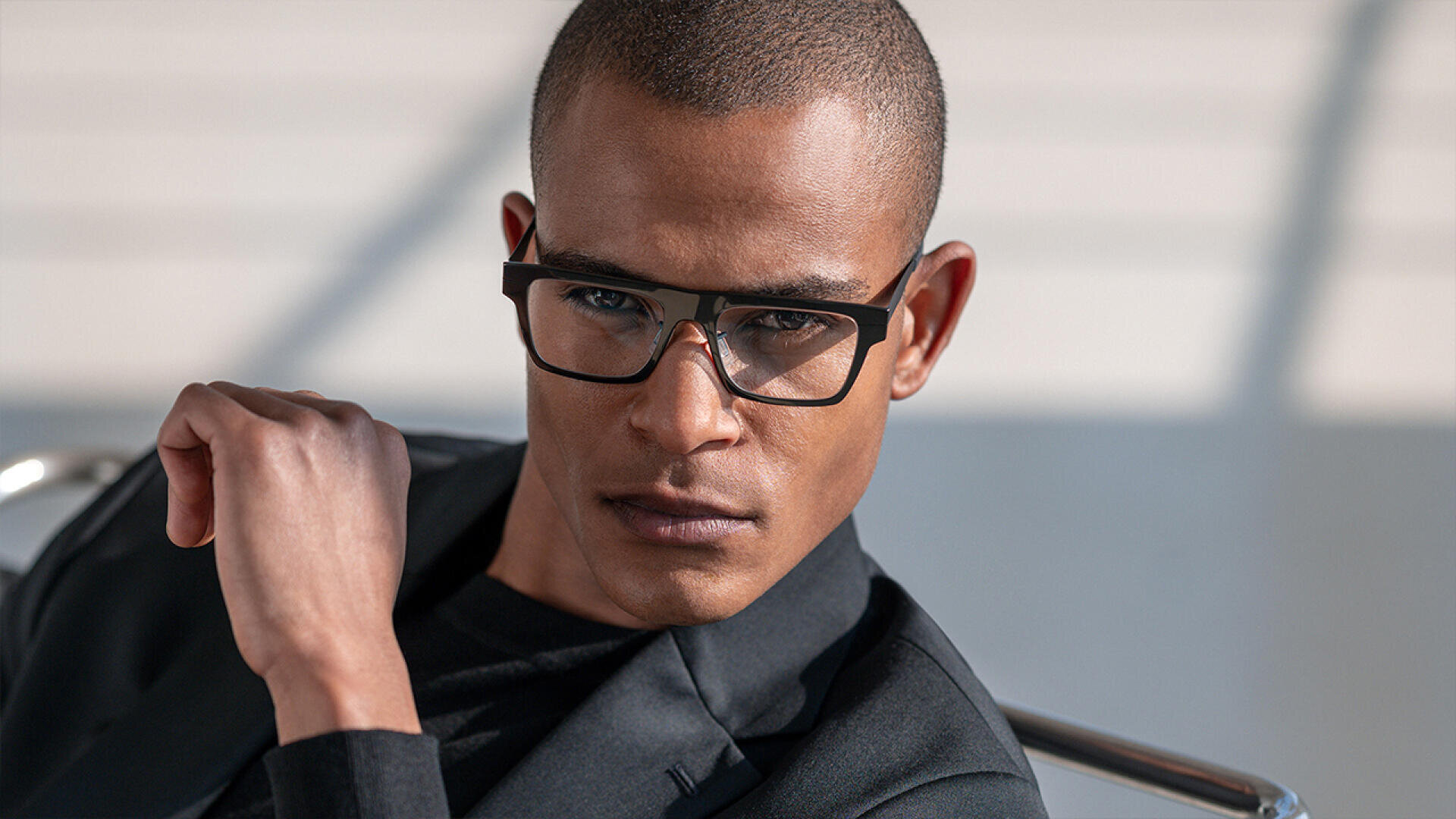 A man wearing rectangular black glasses and a dark blazer looks confidently at the camera while seated, with his arm slightly raised and natural light falling on his face.