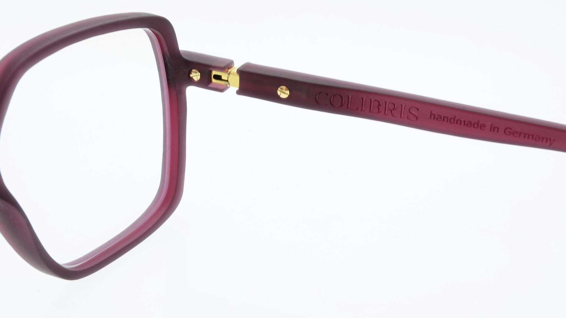 Close-up of the side of a pair of purple eyeglasses; the arm features engraved text reading COLIBRIS handmade in Germany with gold hinge details visible.